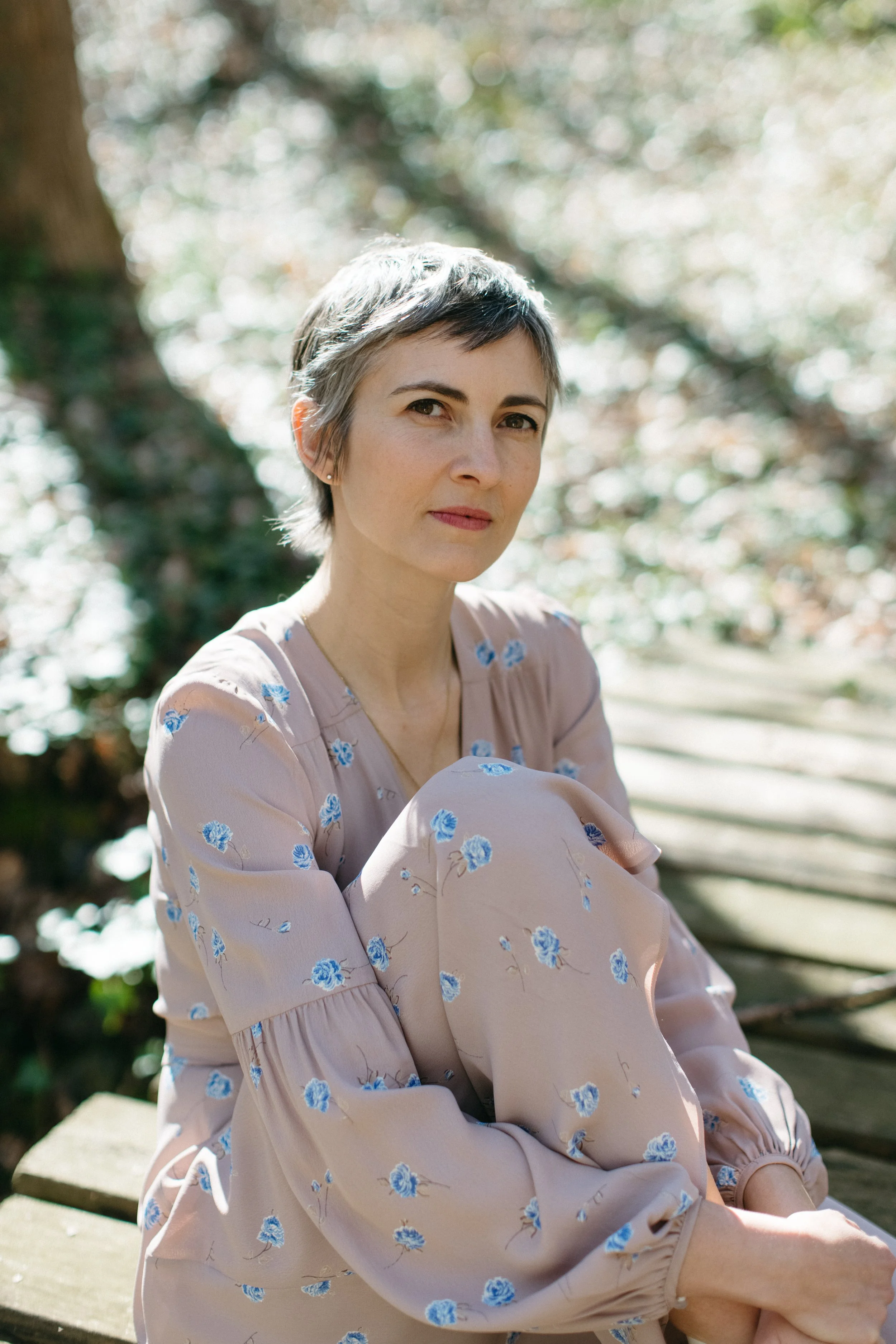 A woman with short hair sitting on a wooden footbridge outdoors, wearing a light pink dress with blue floral patterns, surrounded by blurred trees and foliage in the background.