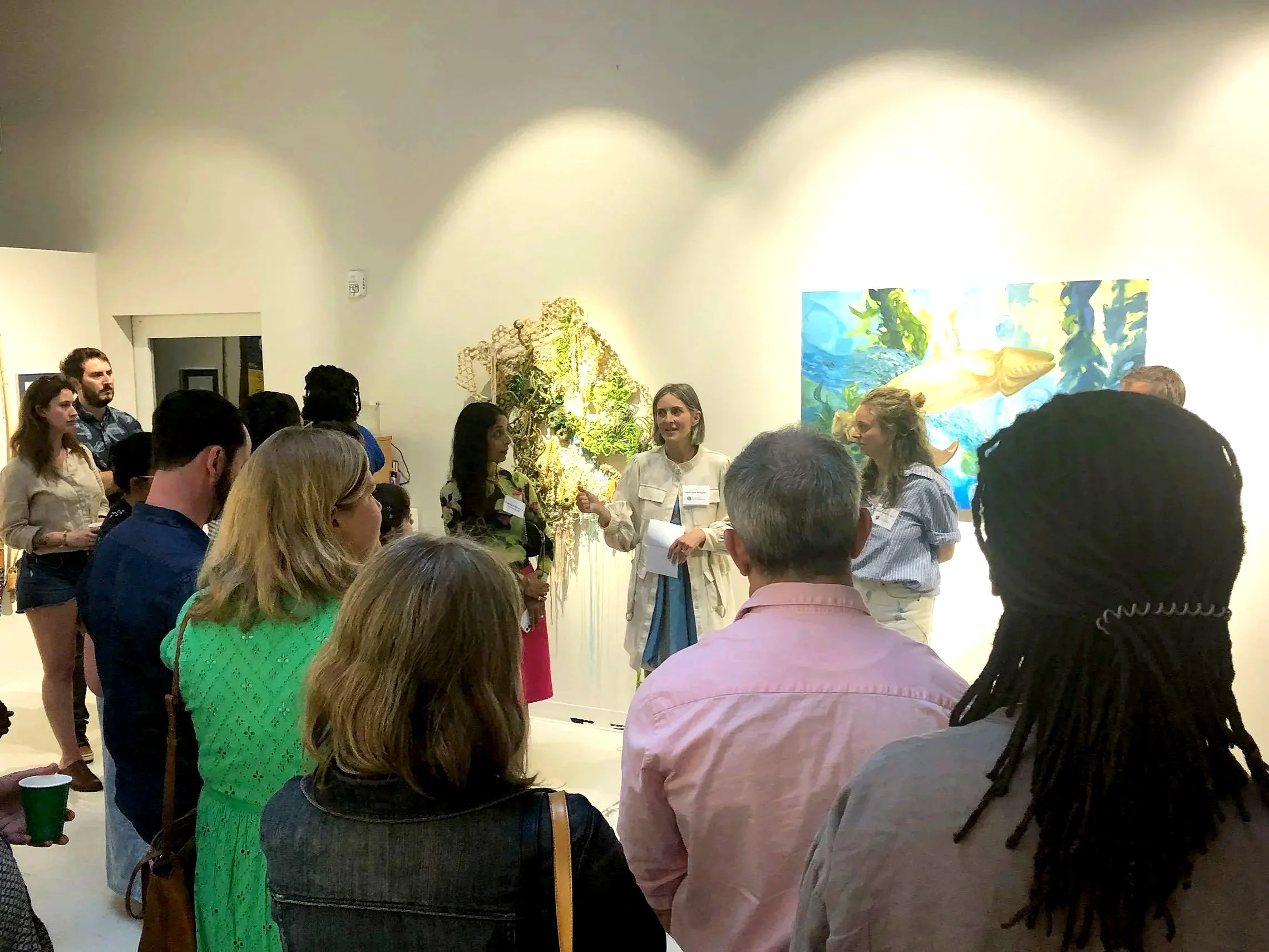 Group of people standing and listening to a woman speaking in an art gallery. The gallery wall behind her has colorful artwork, including a large map and a painting of a fish.