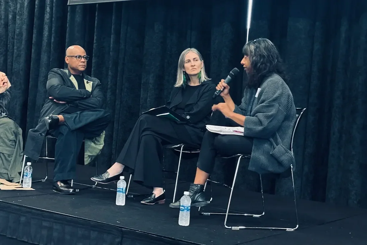 Three people seated on a stage with black curtains in the background. One woman is speaking into a microphone, holding papers, while the other two are listening. Water bottles are on the floor in front of them.
