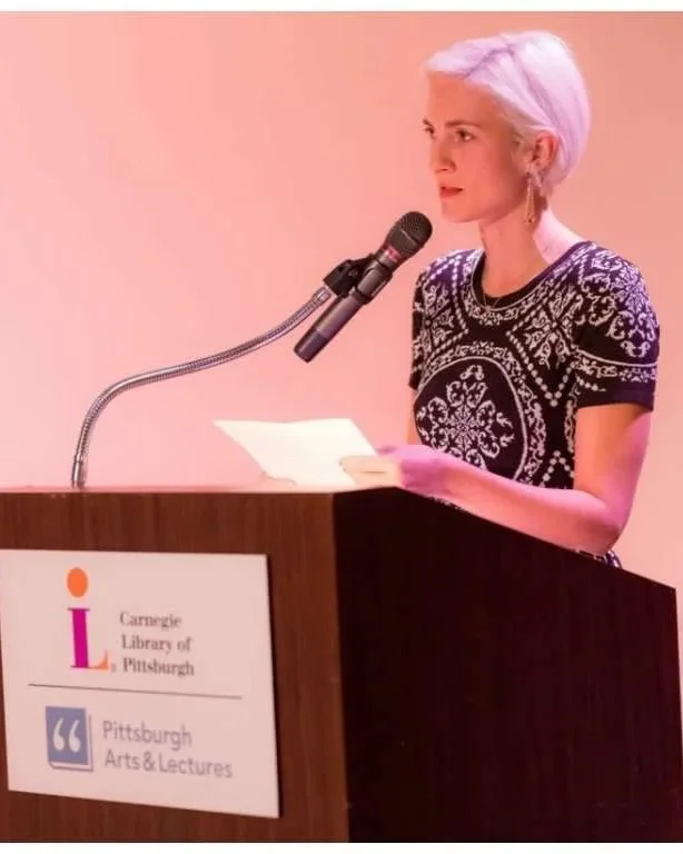 A woman with short white-blonde hair standing at a podium, speaking into a microphone, holding a piece of paper. The podium has a sign for Carnegie Library of Pittsburgh and Pittsburgh Arts & Lectures.