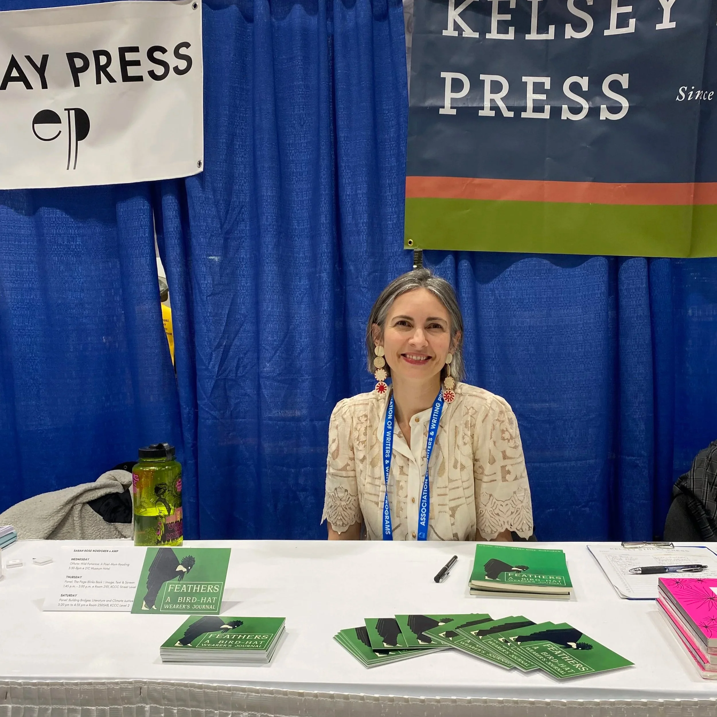 Table with green and black flyers titled "FEATHERS A BIRD-HAT WEARER'S JOURNAL," with stacks of green flyers, pink flyers, and pens, behind blue curtains at an event.