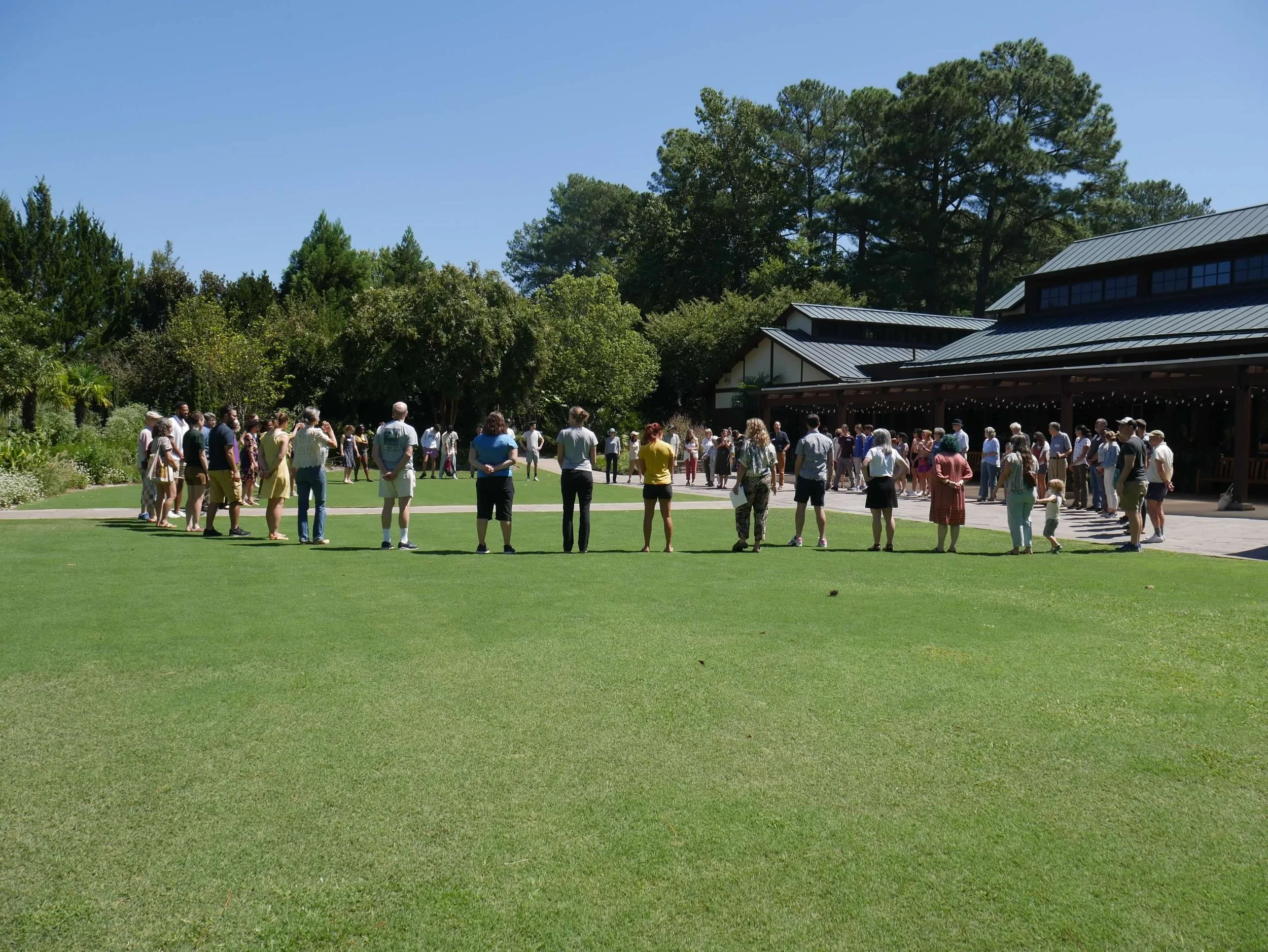 A large group of people gathered outdoors on a lawn area, with trees and a building with a metal roof in the background, on a clear sunny day.
