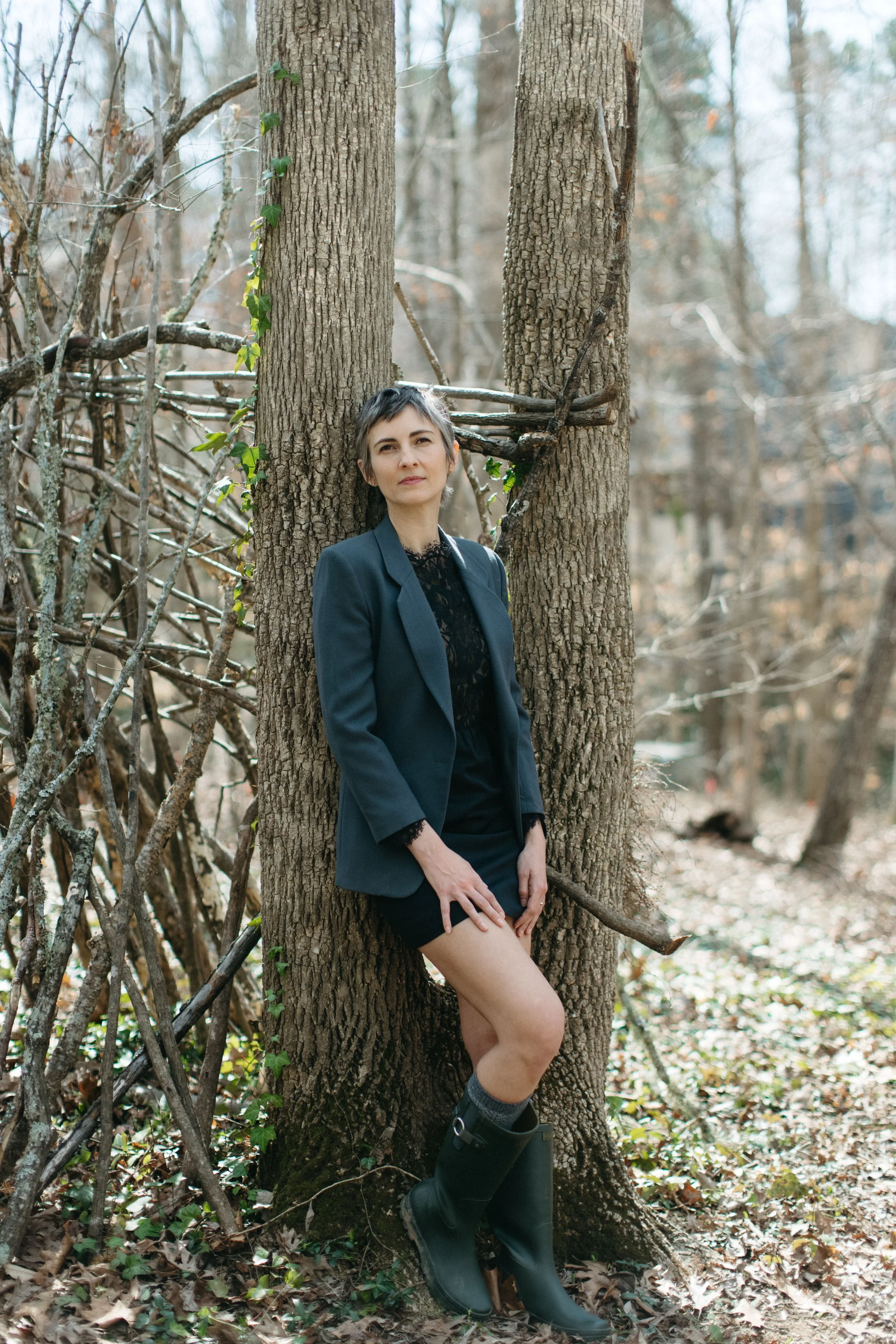 A woman wearing a black lace dress, blazer, and knee-high rain boots standing in a forest with her back against a tree. She has short dark hair and is looking into the distance.