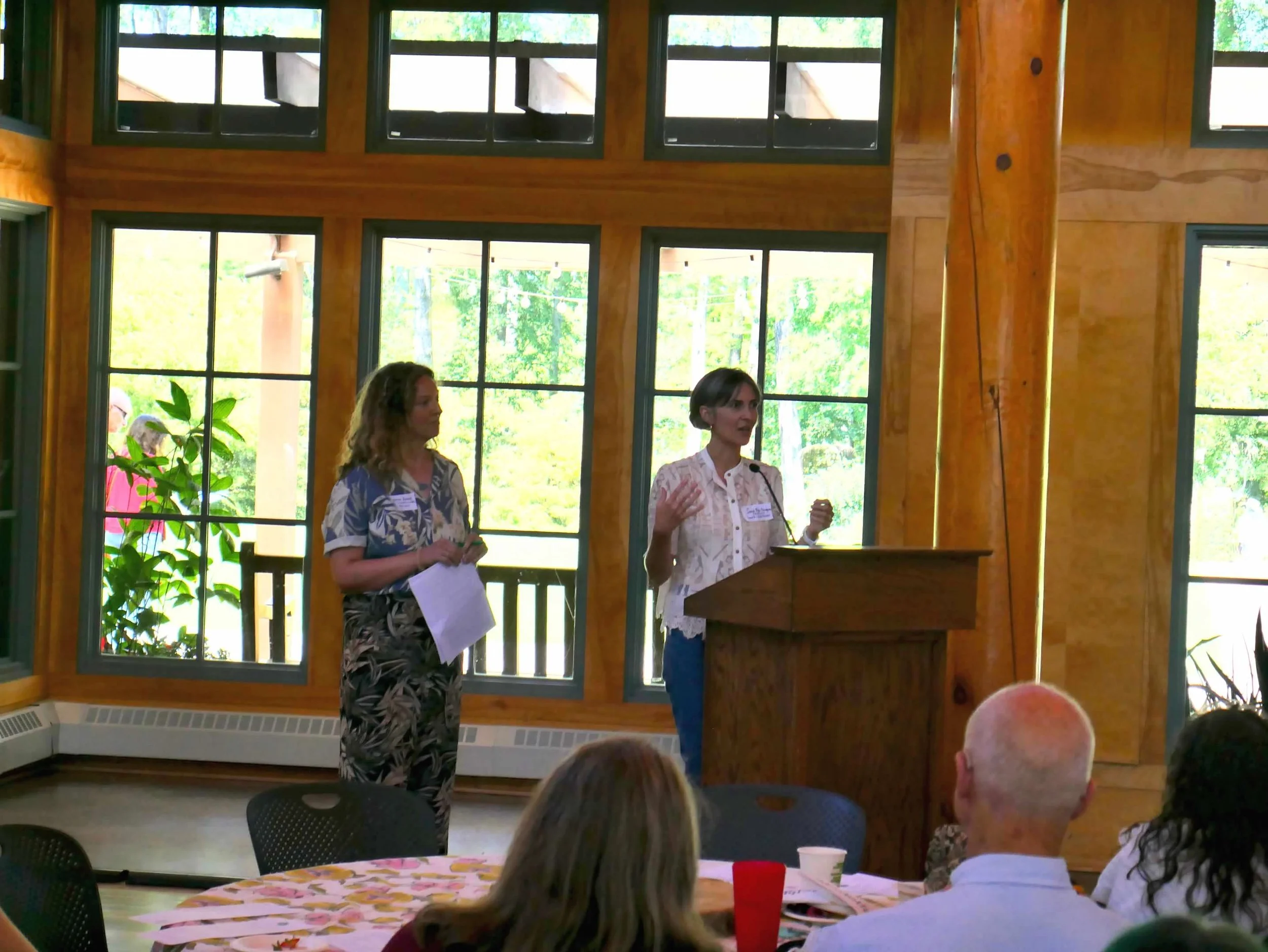 Two women speaking at a wooden podium in a room with large windows and natural light. Several people are seated at tables facing them.