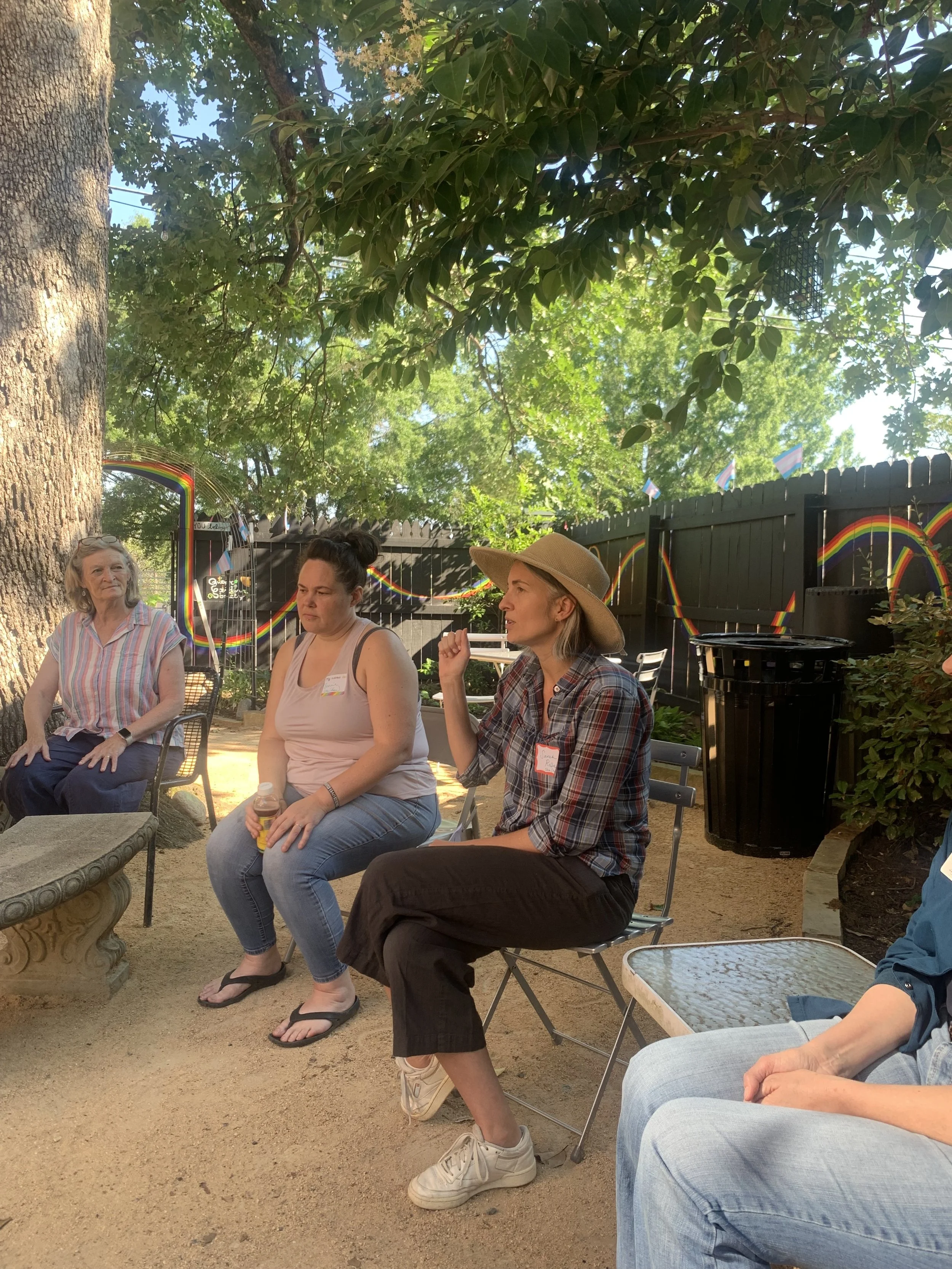 Group of people sitting outdoors in a garden, engaged in a conversation, with a large tree, a black fence decorated with rainbow flags, and rainbow-themed decorations in the background.