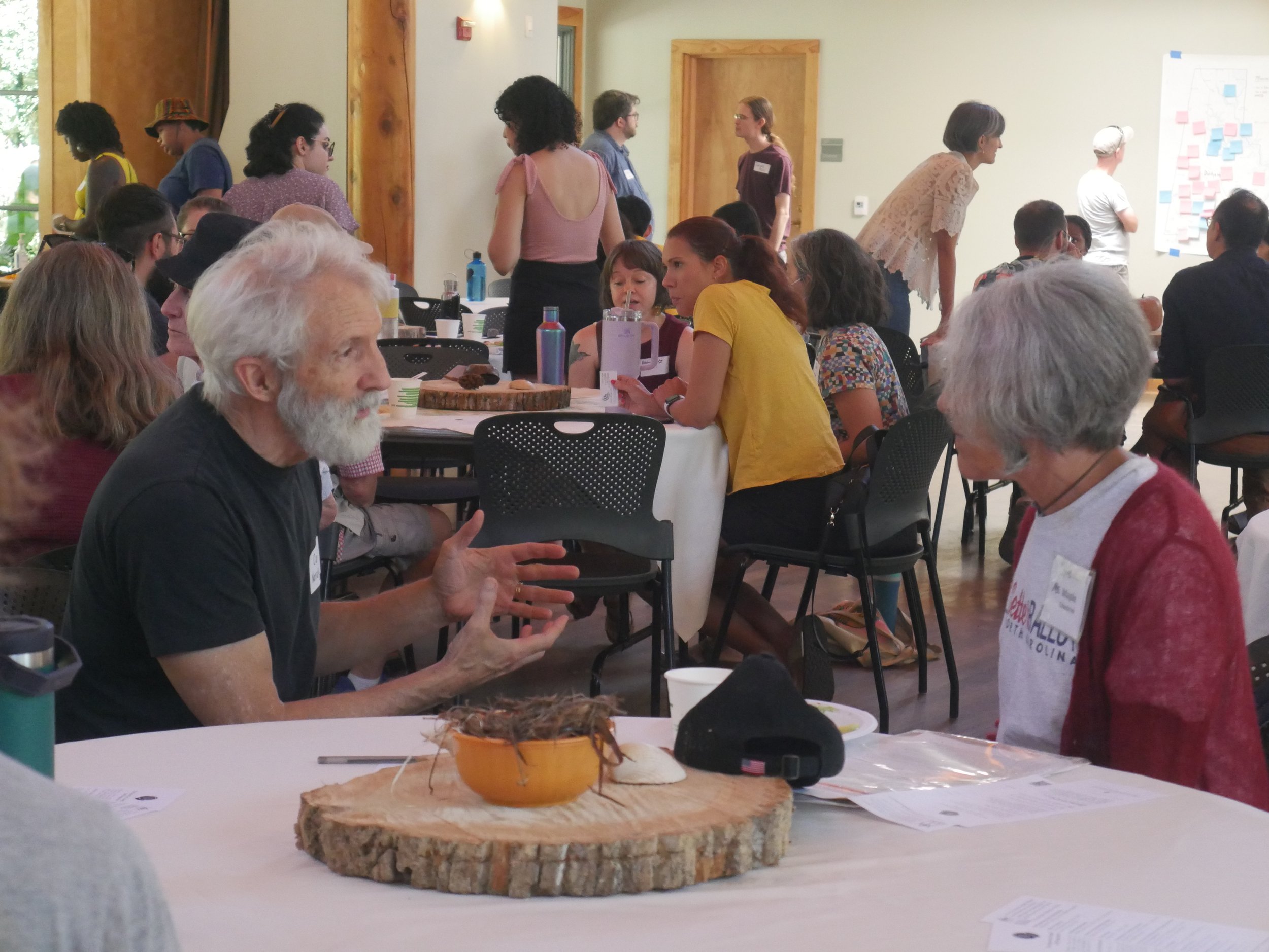 A group of people gathered in a room, some seated at tables and others standing, engaging in conversations during a social or community event. There is a wooden slab with a small orange container on it in the foreground.