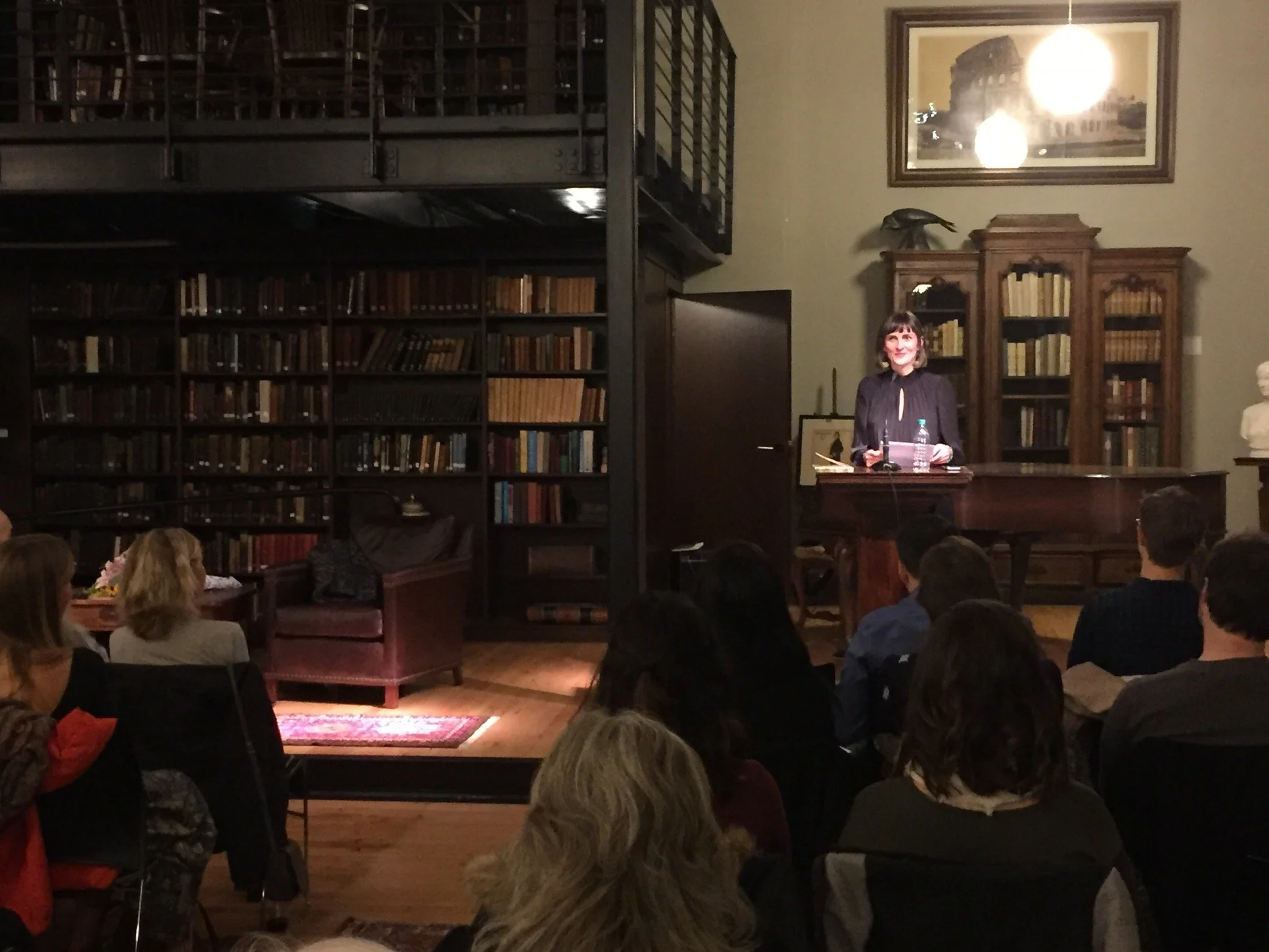 A woman with short dark hair, wearing a black top, stands behind a wooden podium, smiling during a presentation or speech in a room with tall bookshelves filled with books. The audience is seated in front of her, and there is a framed picture of the 