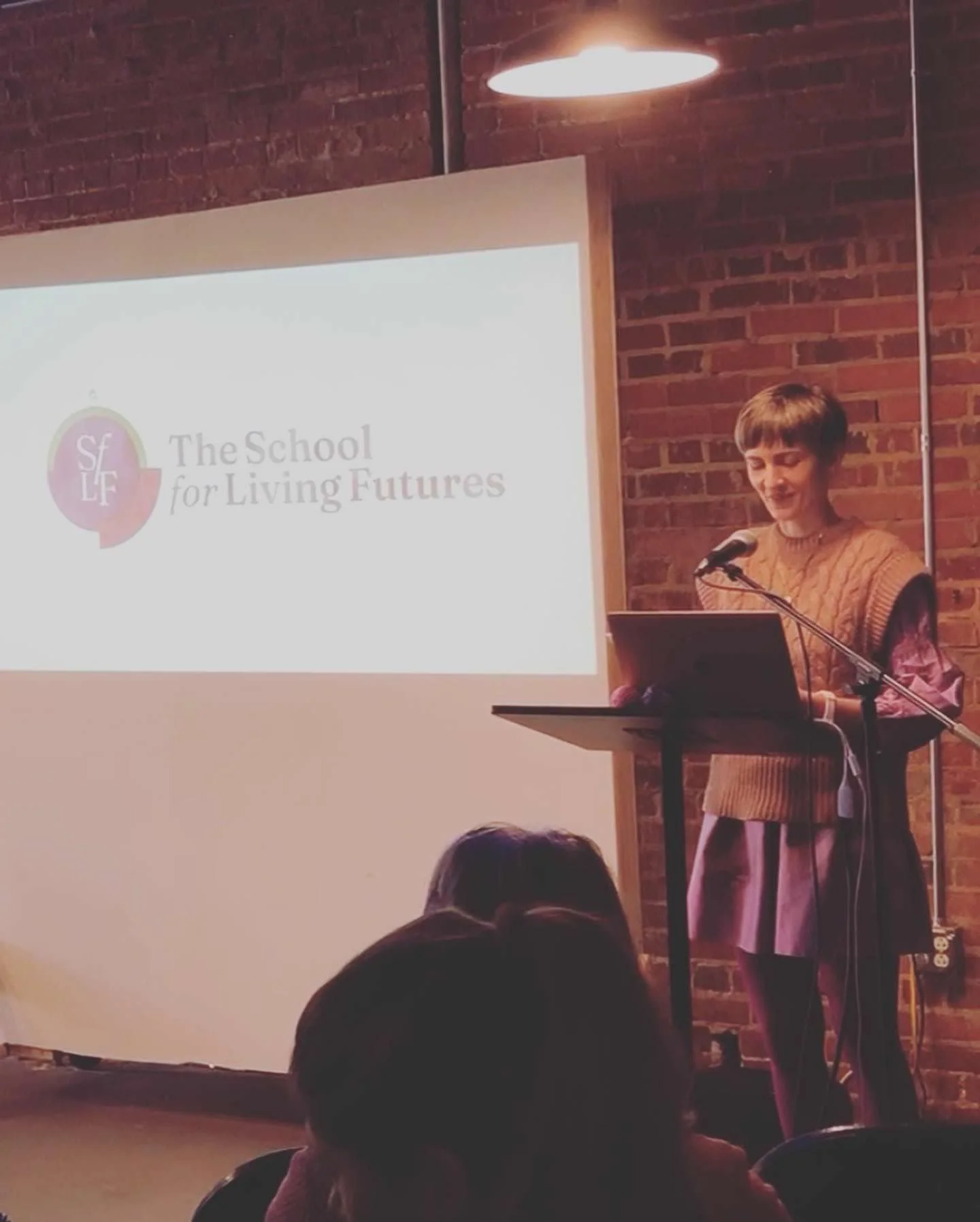 A woman standing at a podium with a microphone, reading or speaking during a presentation in a room with brick walls. A projector screen displays a logo and text that says, "The School for Living Futures."