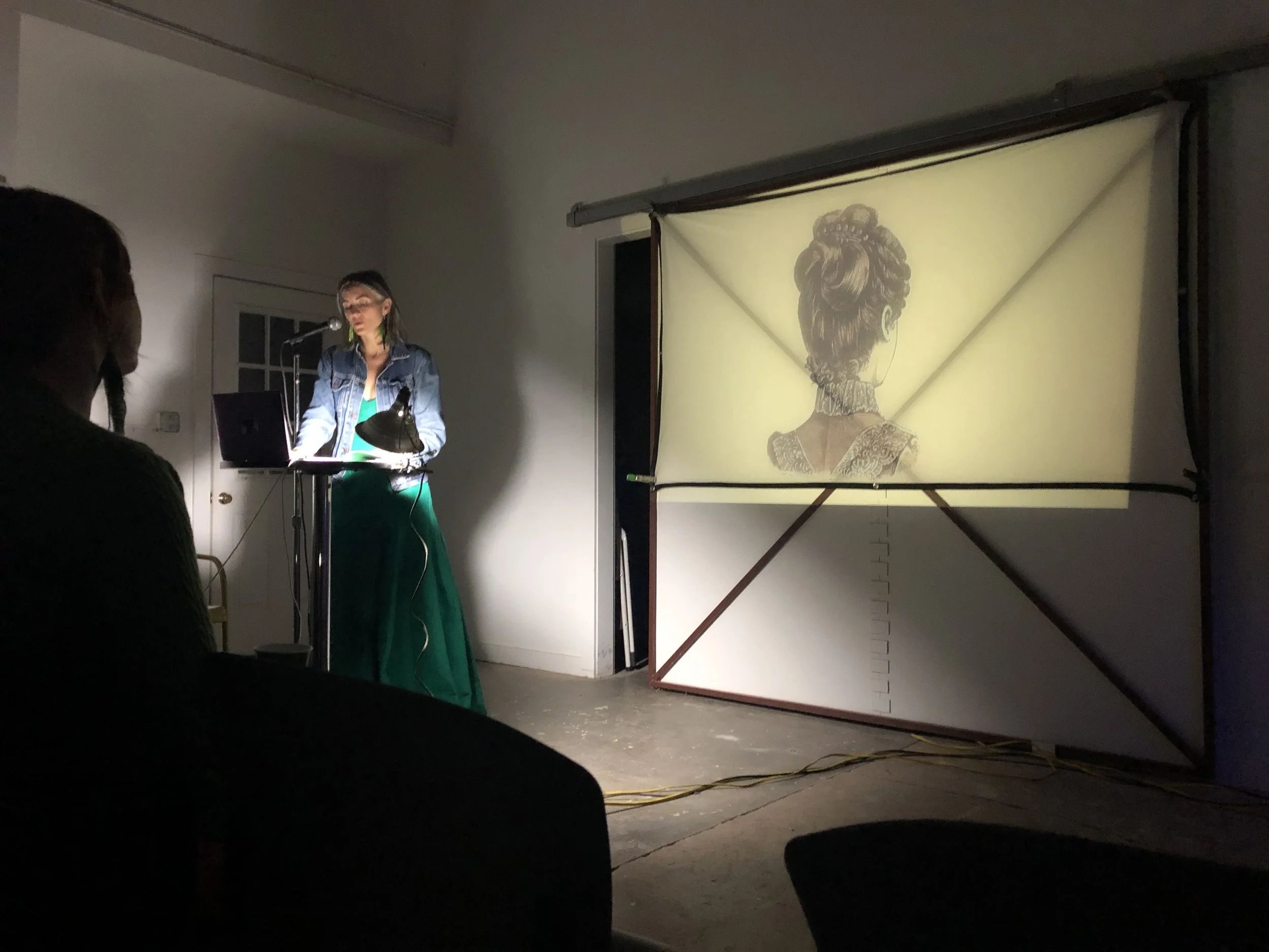A woman is standing at a podium with a microphone, reading or speaking, indoors. To her right, there is a large projection screen displaying an artistic illustration of a woman with an elaborate hairstyle and ornate dress, viewed from the back.