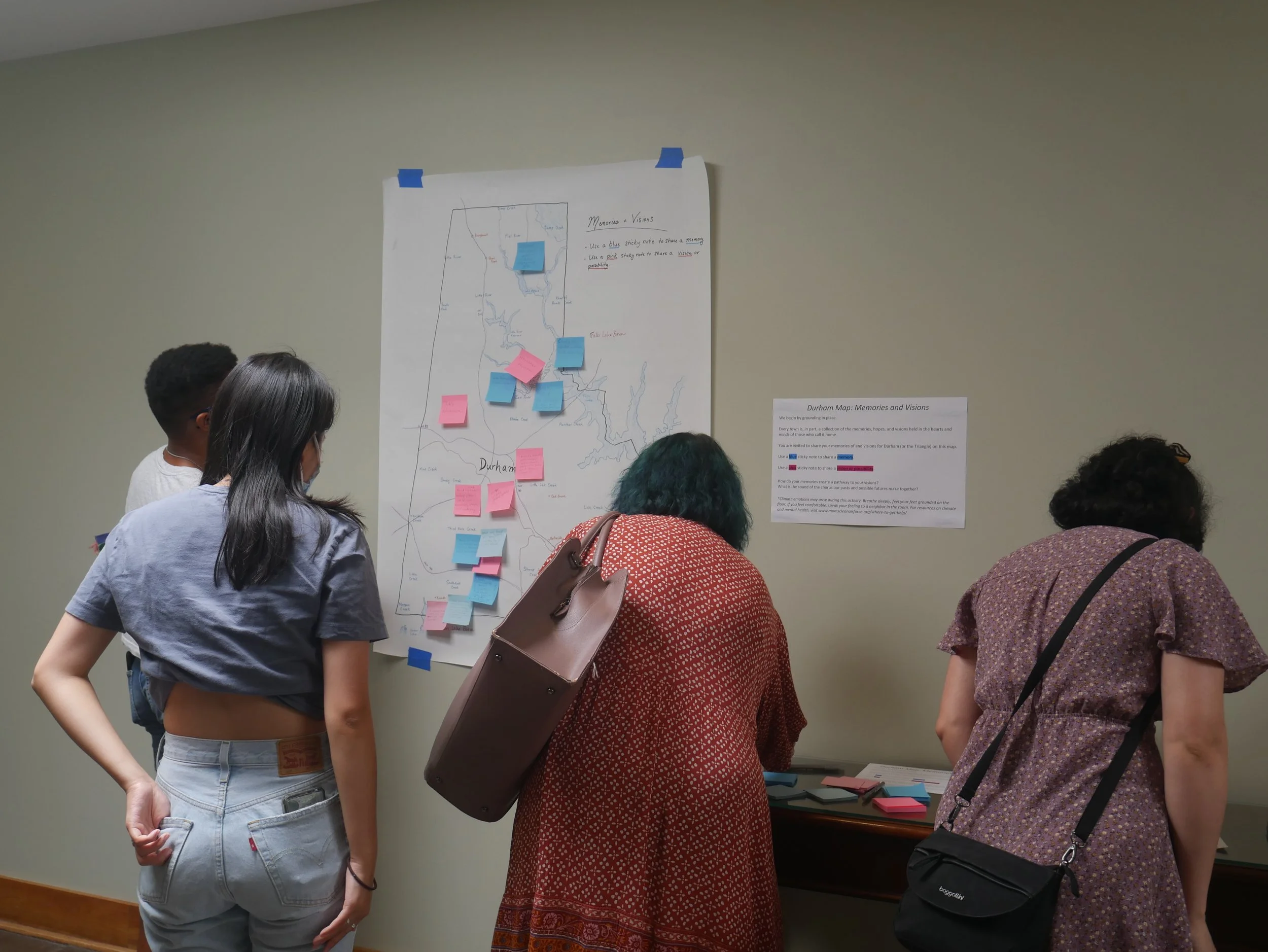 People looking at a large poster board with notes and a map, discussing. Several pink and blue sticky notes are attached to the map on the board.