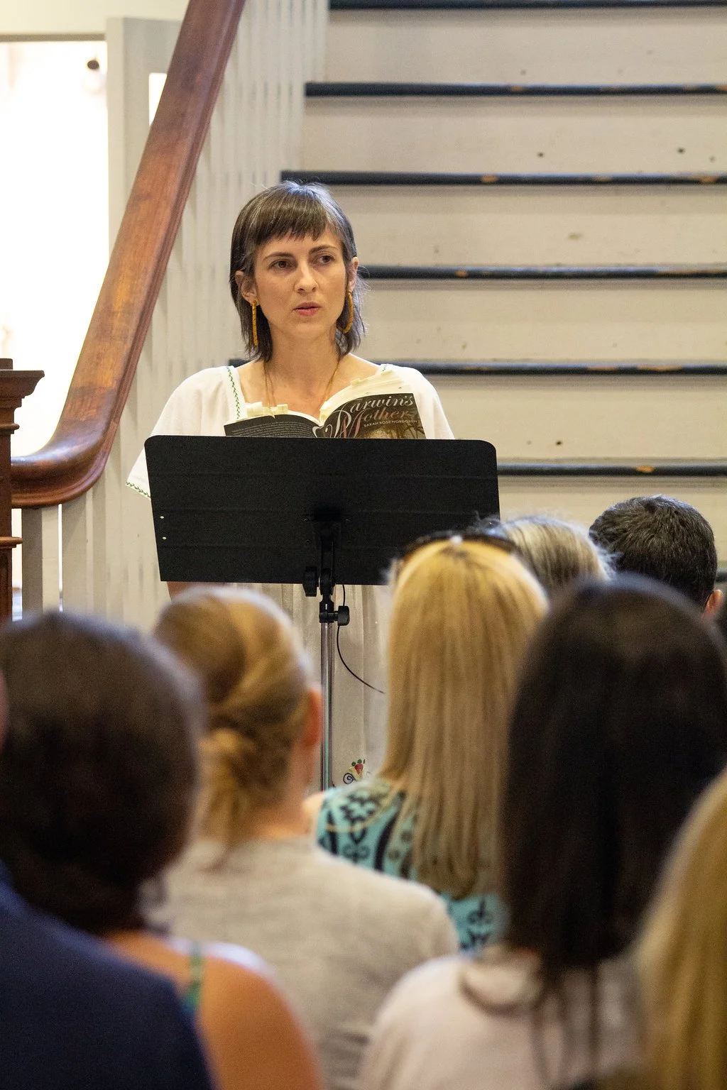 A woman standing behind a music stand, holding a book, speaking to an audience in a classroom or lecture hall setting.