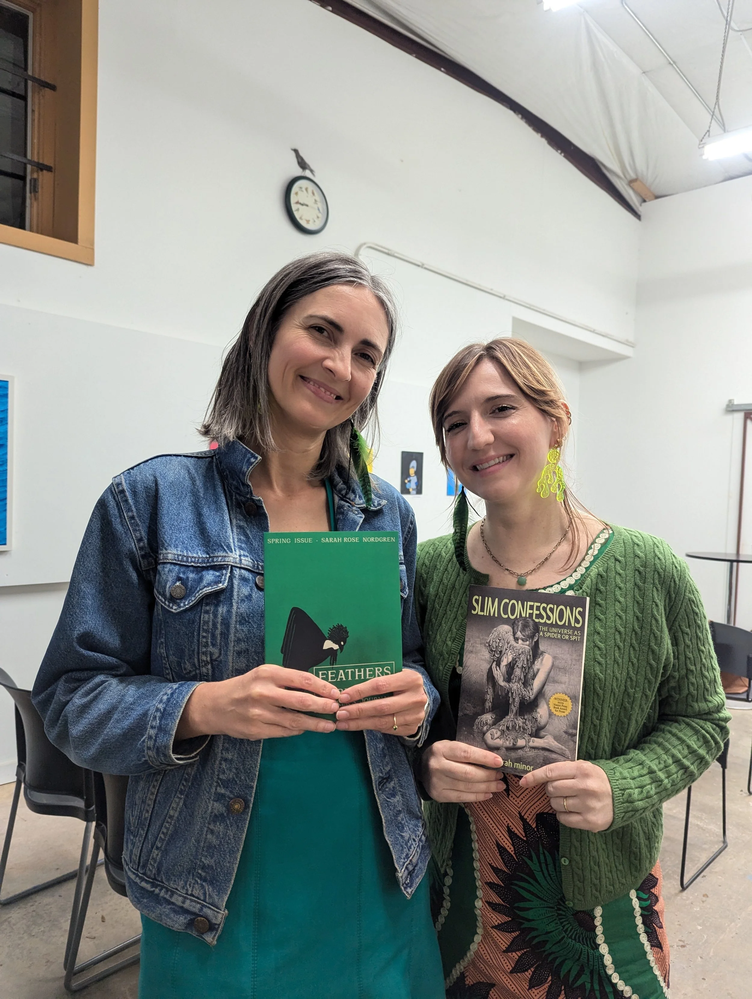 Two women smiling and holding books at an indoor event. The woman on the left is wearing a denim jacket, holding a book titled 'Feathers.' The woman on the right is wearing a green sweater and large yellow earrings, holding a book titled 'Slim Confes