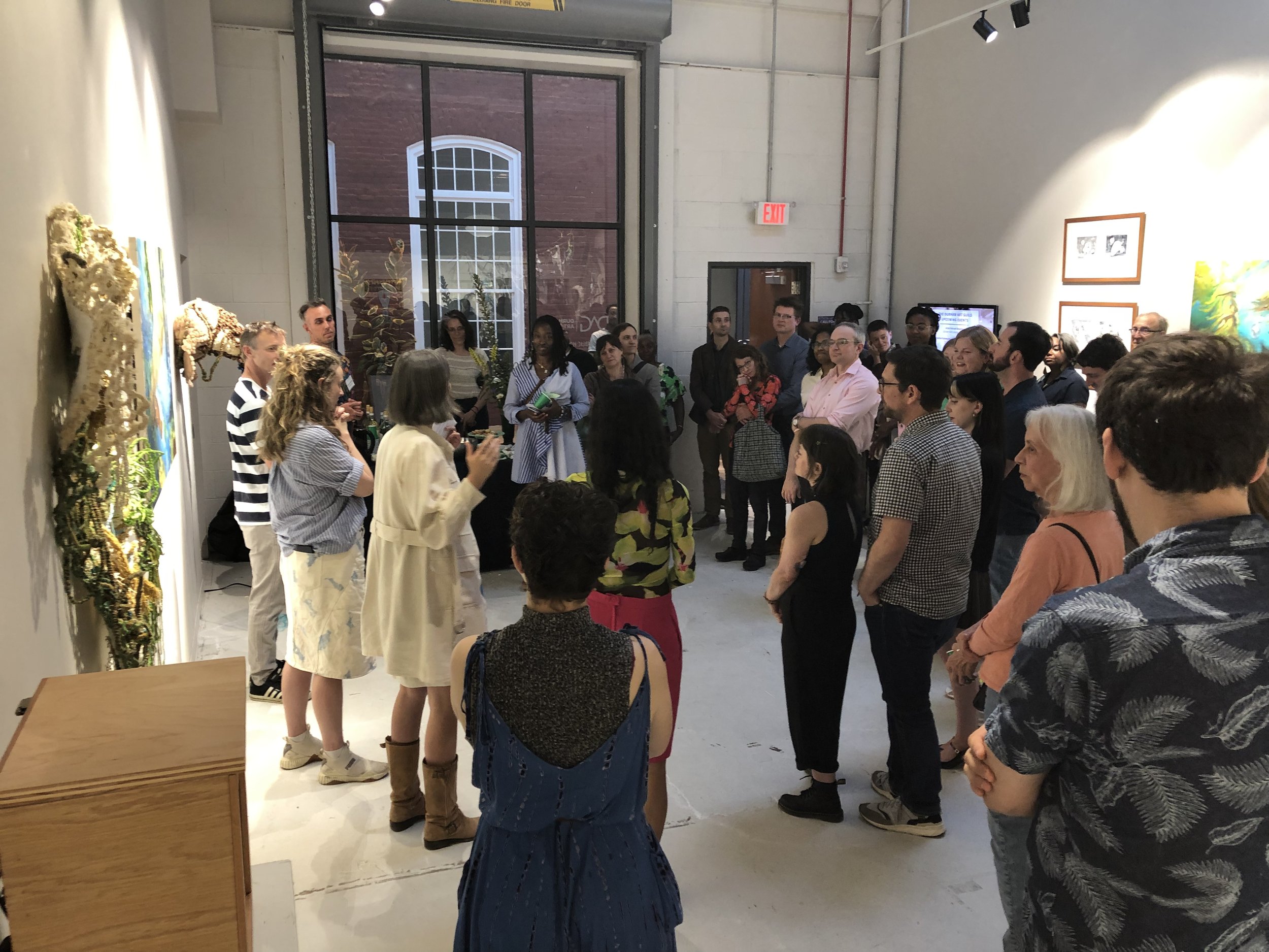 A group of people gathered in an art gallery, listening to a woman speaking. Artworks are displayed on the walls, and there's a large window in the background showing a brick building outside.