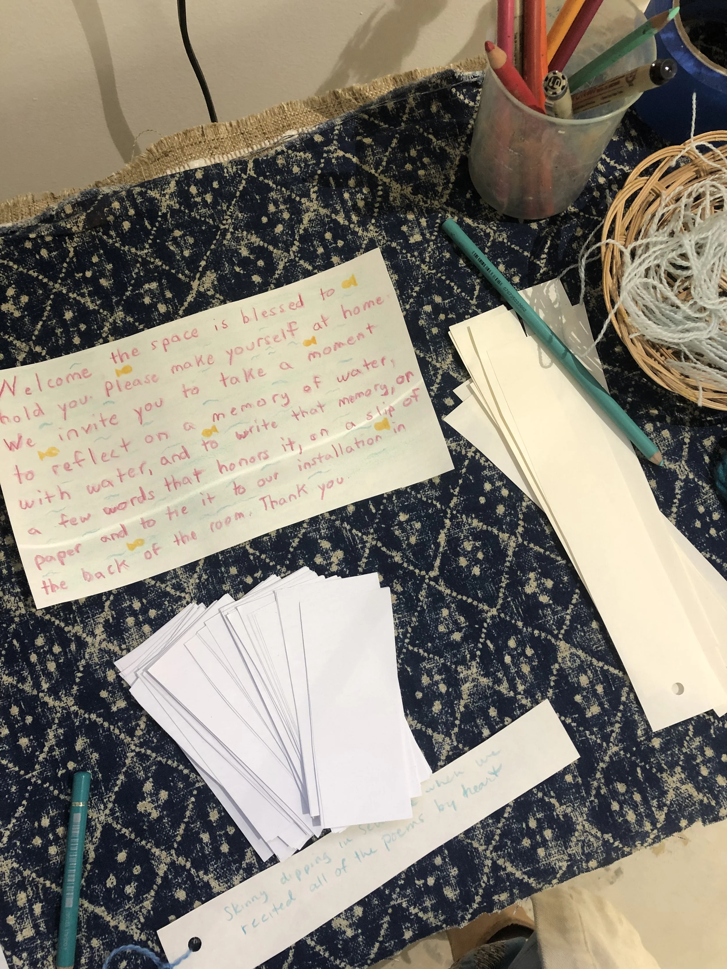 A handwritten note on white paper, surrounded by markers, paper, and string on a dark patterned table.