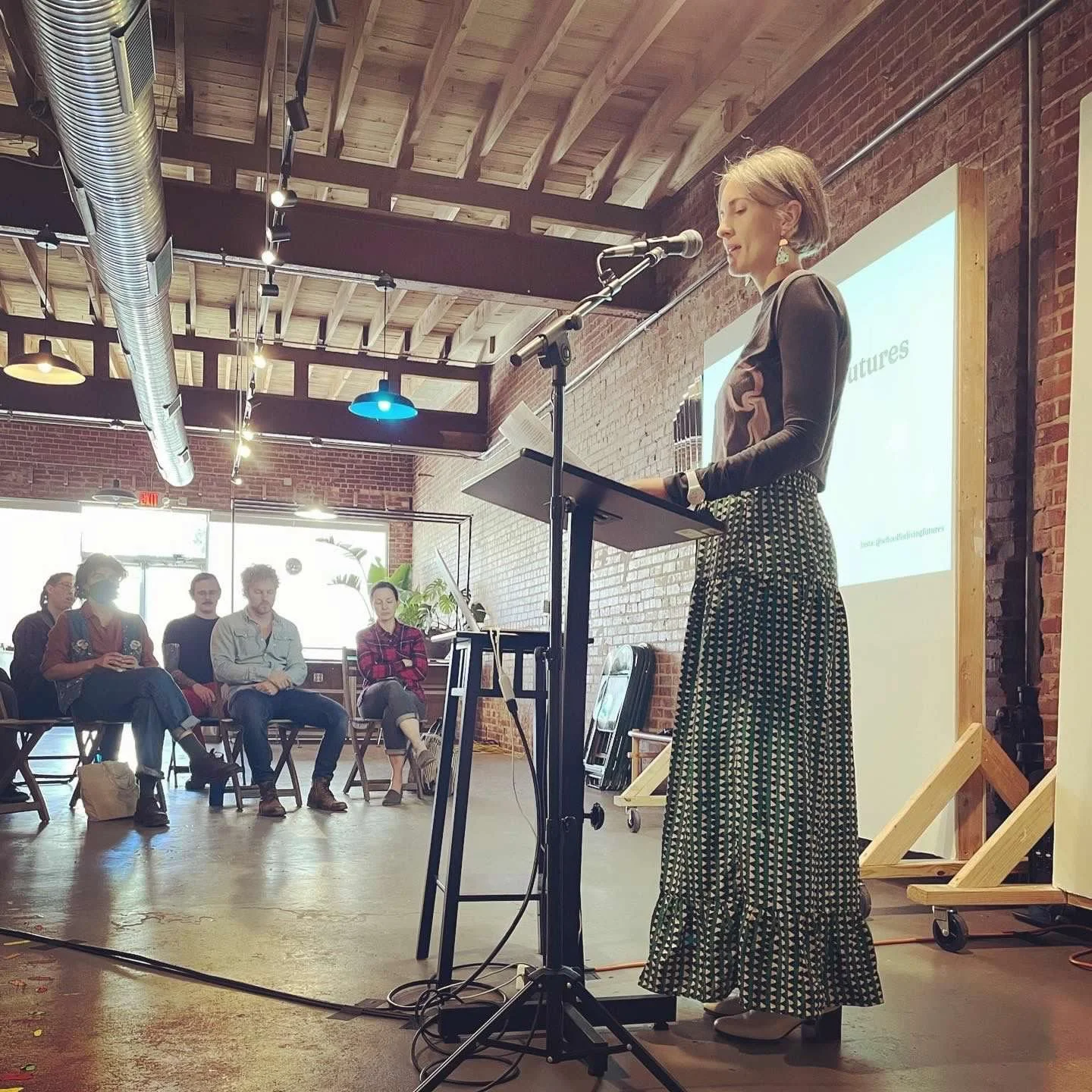 A woman standing at a podium giving a presentation to an audience in a modern, industrial-style room with exposed brick walls and wooden ceiling beams.