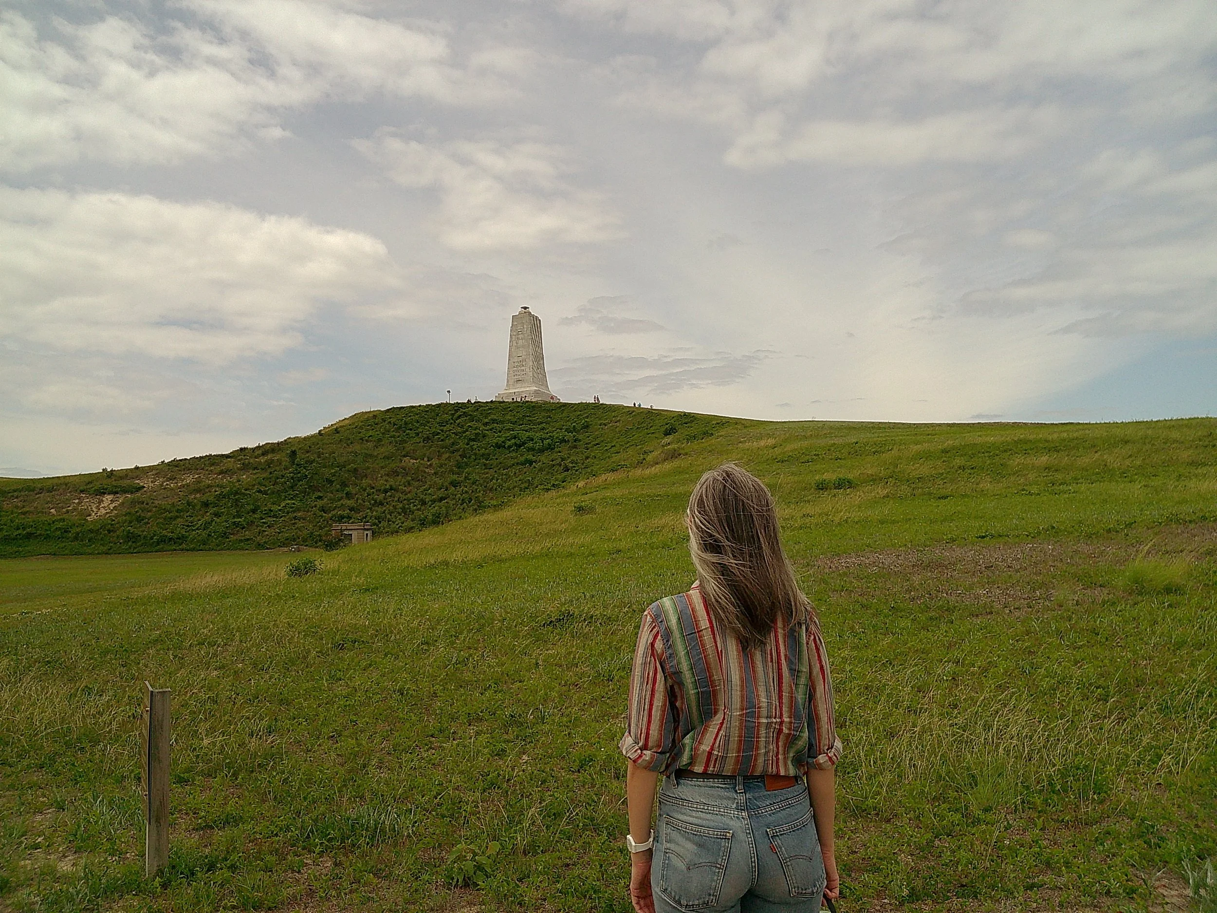 A woman with long hair wearing a plaid shirt and jeans stands outdoors on a grassy hill, looking at a stone monument  with a cloudy sky overhead.