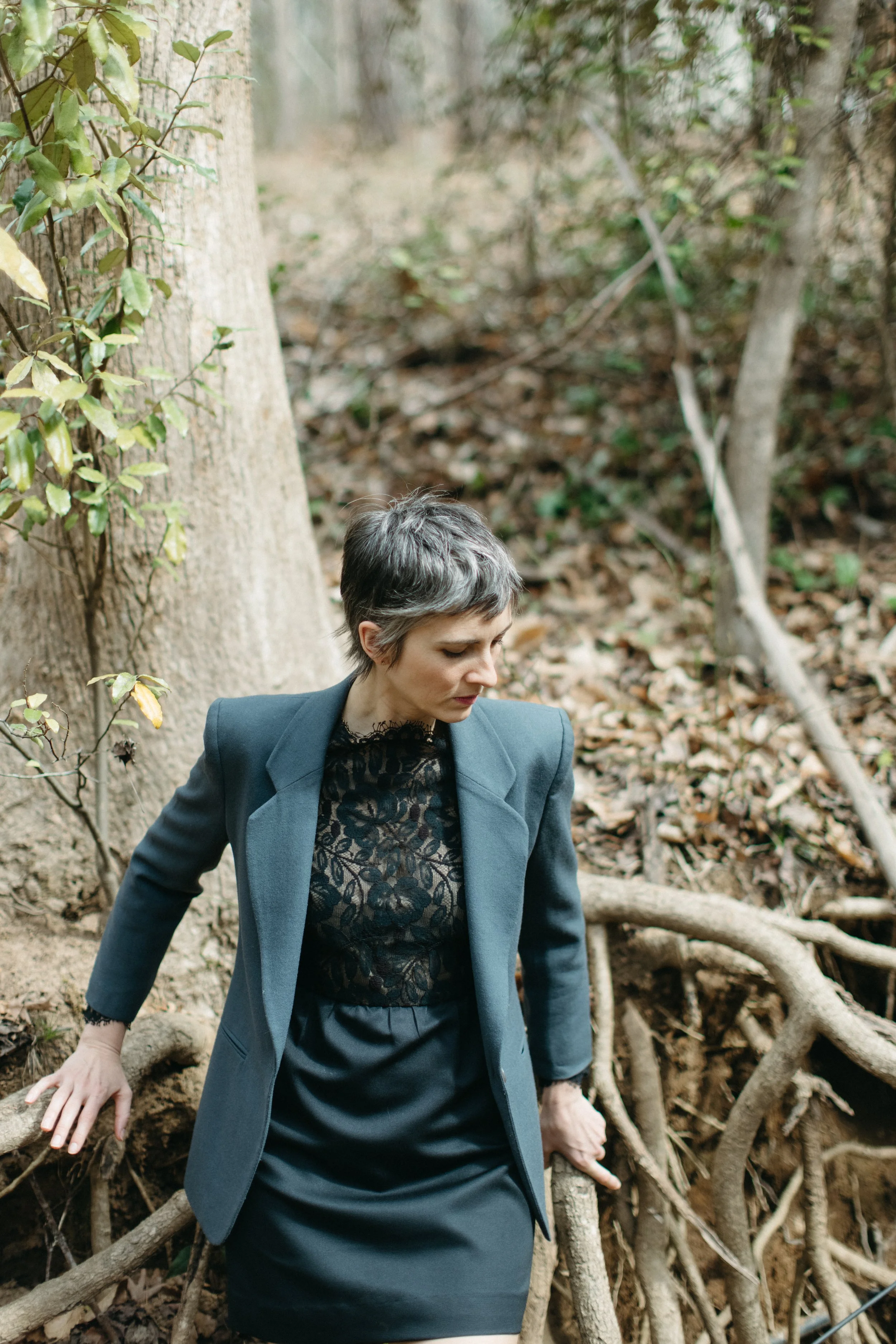 A woman with short, dark hair wearing a dark-colored lace top and a blue blazer, standing in a wooded area with tree roots and fallen leaves.