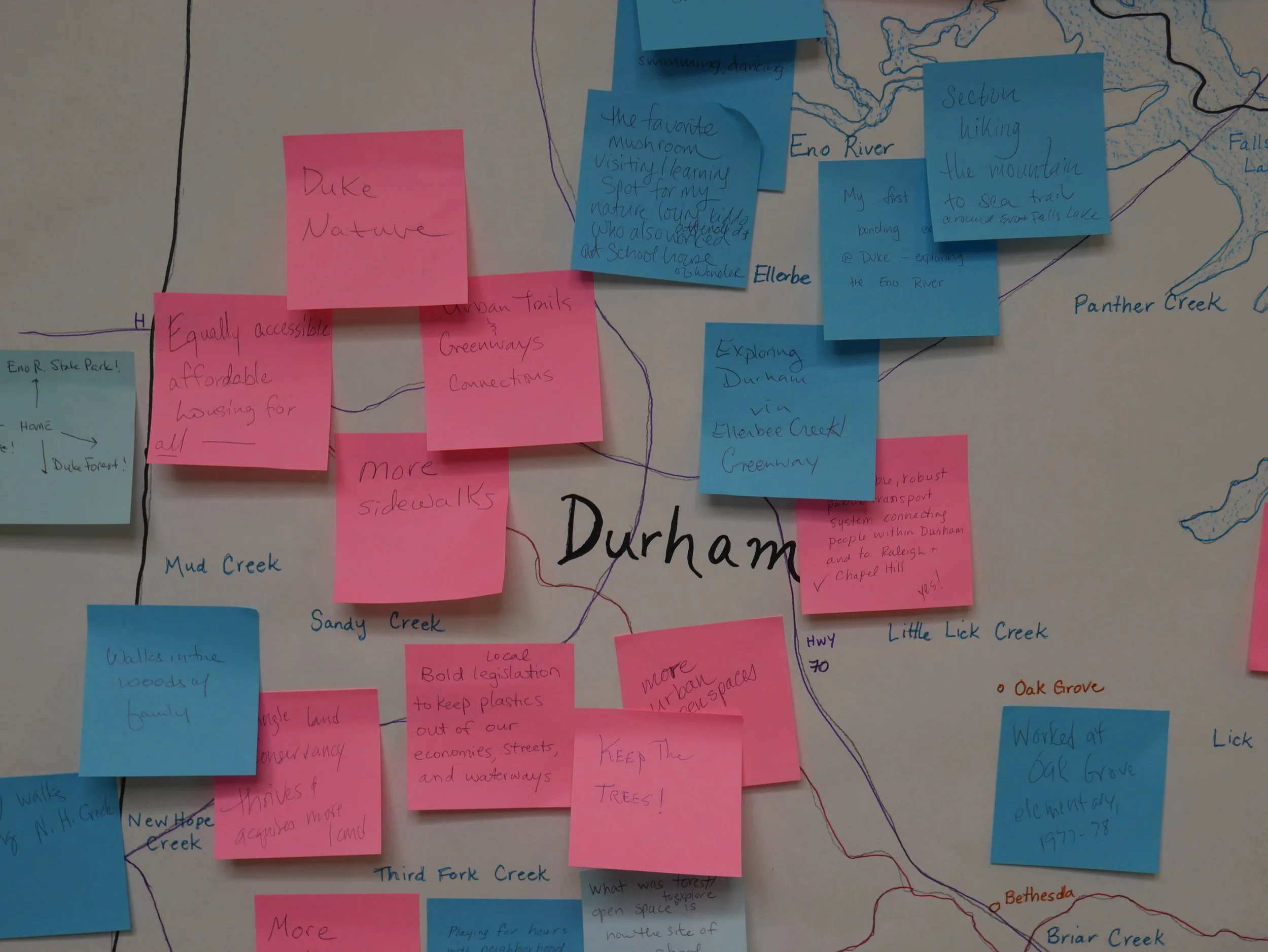A close-up photo of a wall with social notes on colored sticky notes, including blue and pink ones, and a map with handwritten notes about the Durham area and nearby streams like Eno River, Panther Creek, and Little Lick Creek.