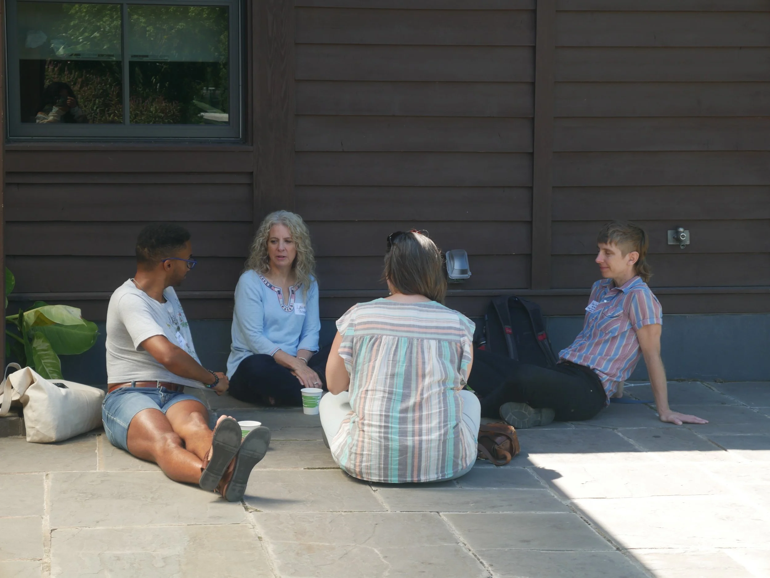 Group of five people sitting outside on a concrete surface, engaged in conversation, with some holding cups, against a wooden wall with a window