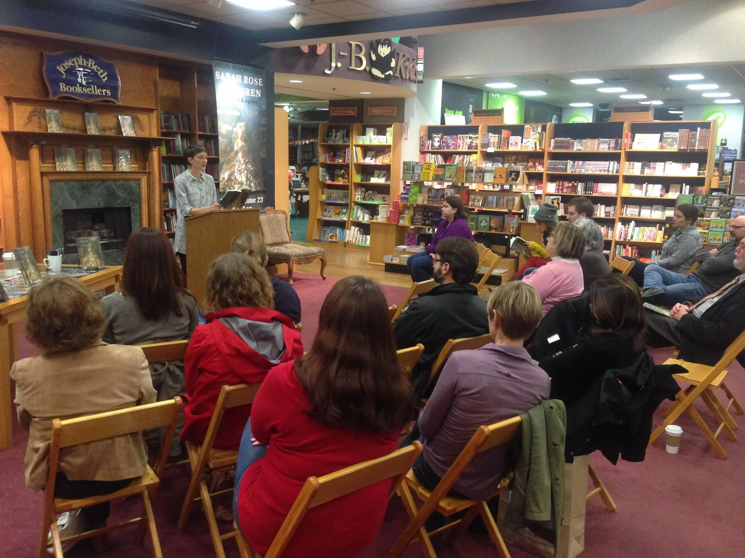 A crowded bookstore with people sitting in chairs and listening to a woman speaking at a podium. Bookshelves filled with books surround the area, and there are signs and posters, including one for Sarah Rose Nordgren.