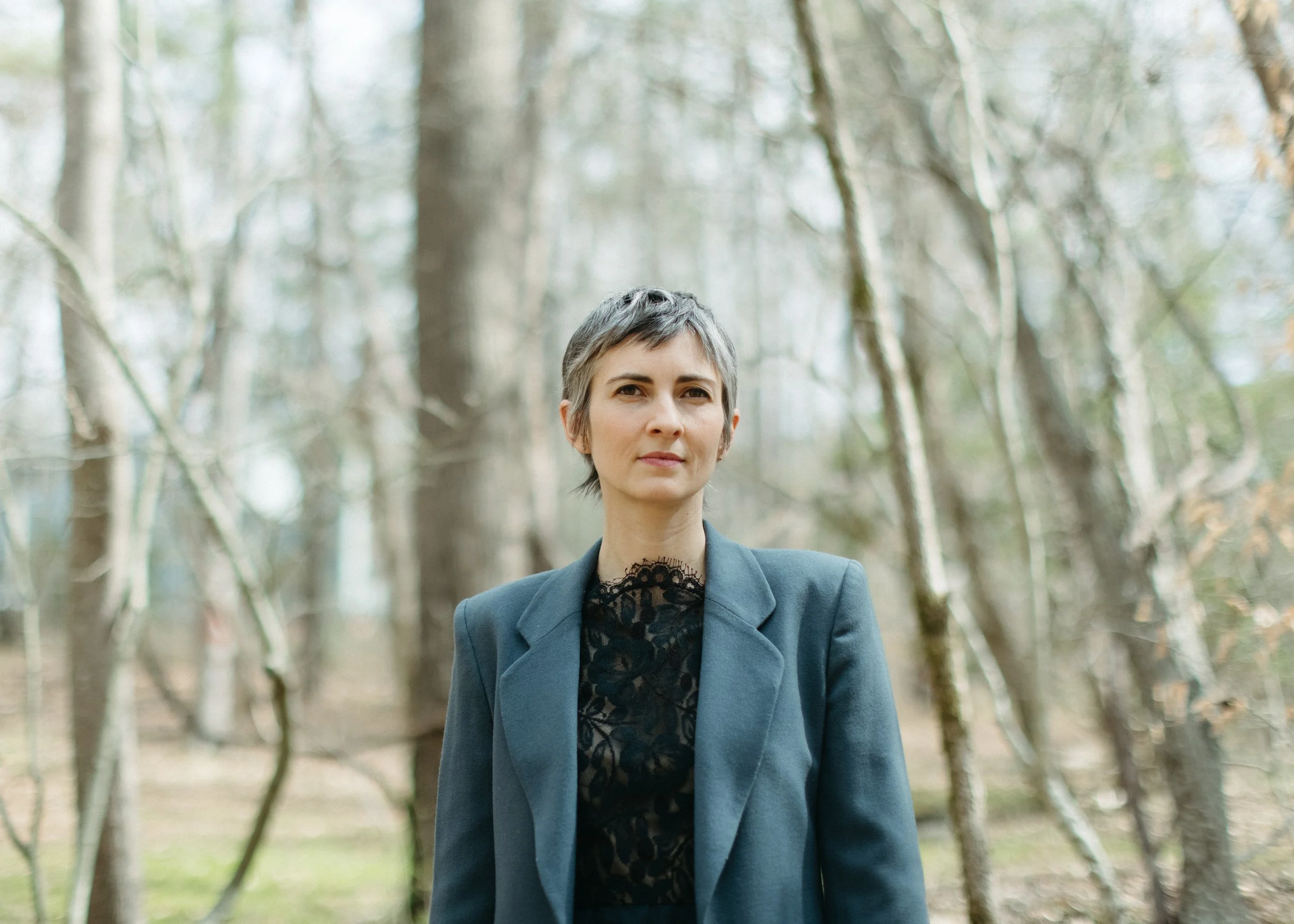 A woman with short hair standing outdoors in a forest during daytime, wearing a gray blazer and black lace top, with a neutral expression.
