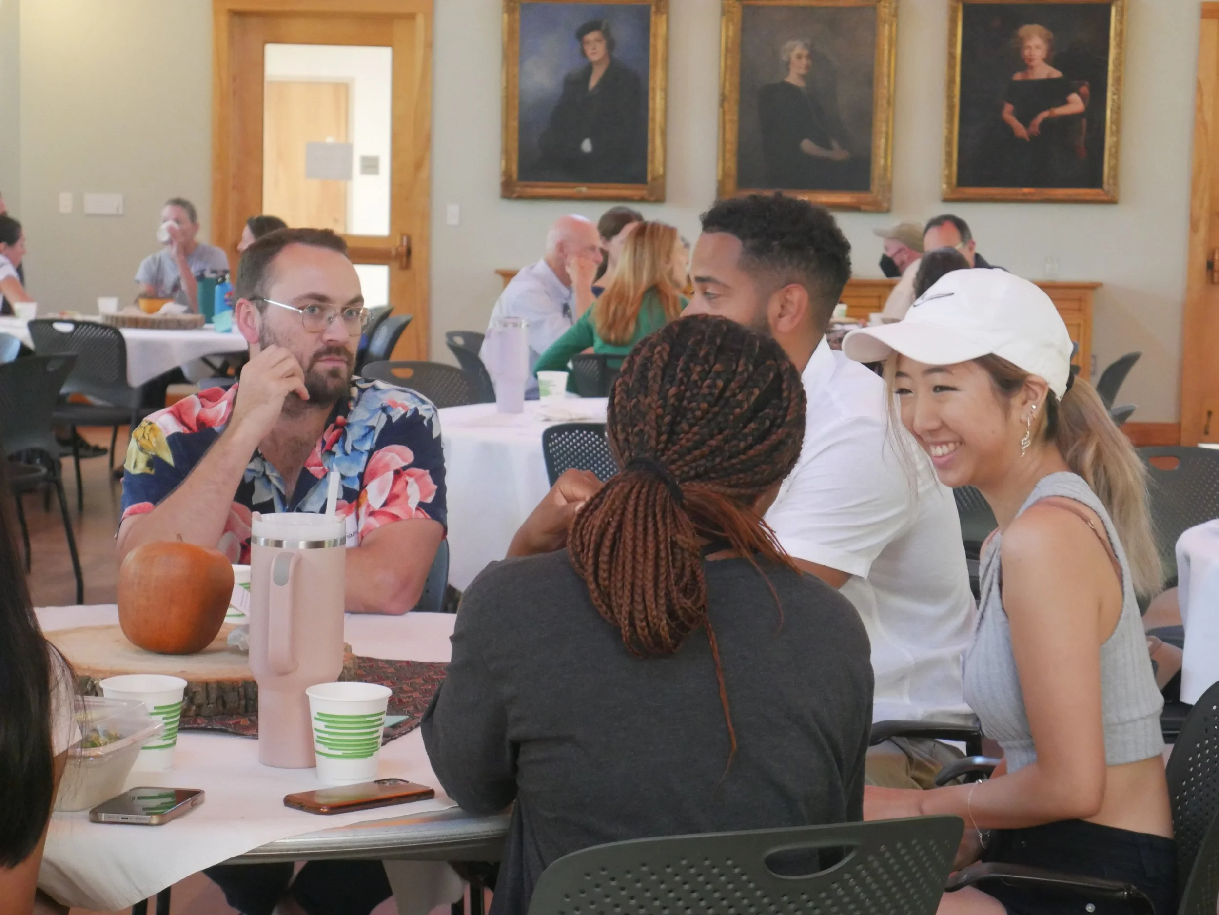 People sitting around a table at an indoor gathering, with paintings hanging on the wall behind them.