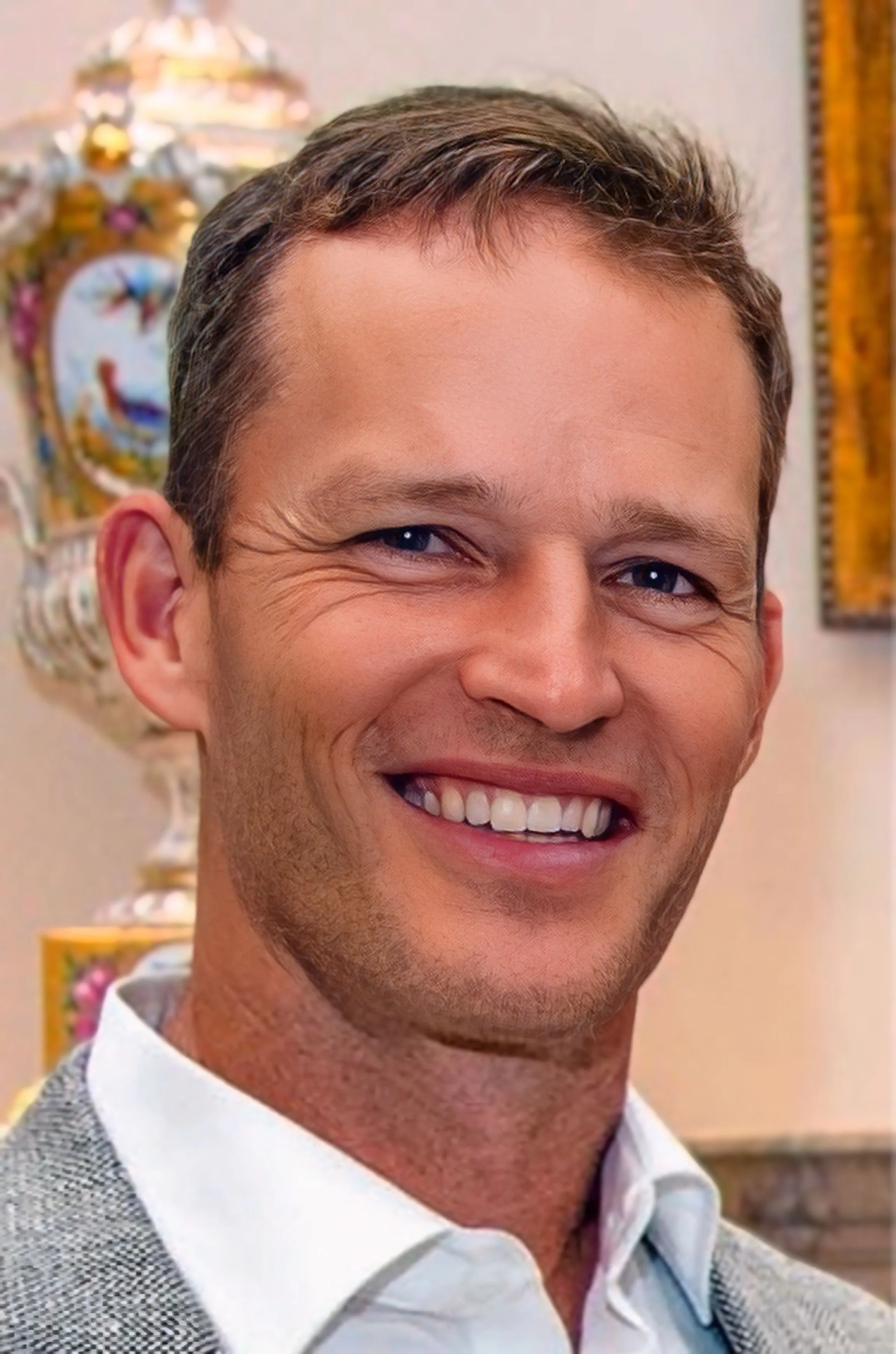 Close-up of smiling man with short brown hair, dressed in a white shirt and gray checked blazer, with colorful decorative objects in the background.