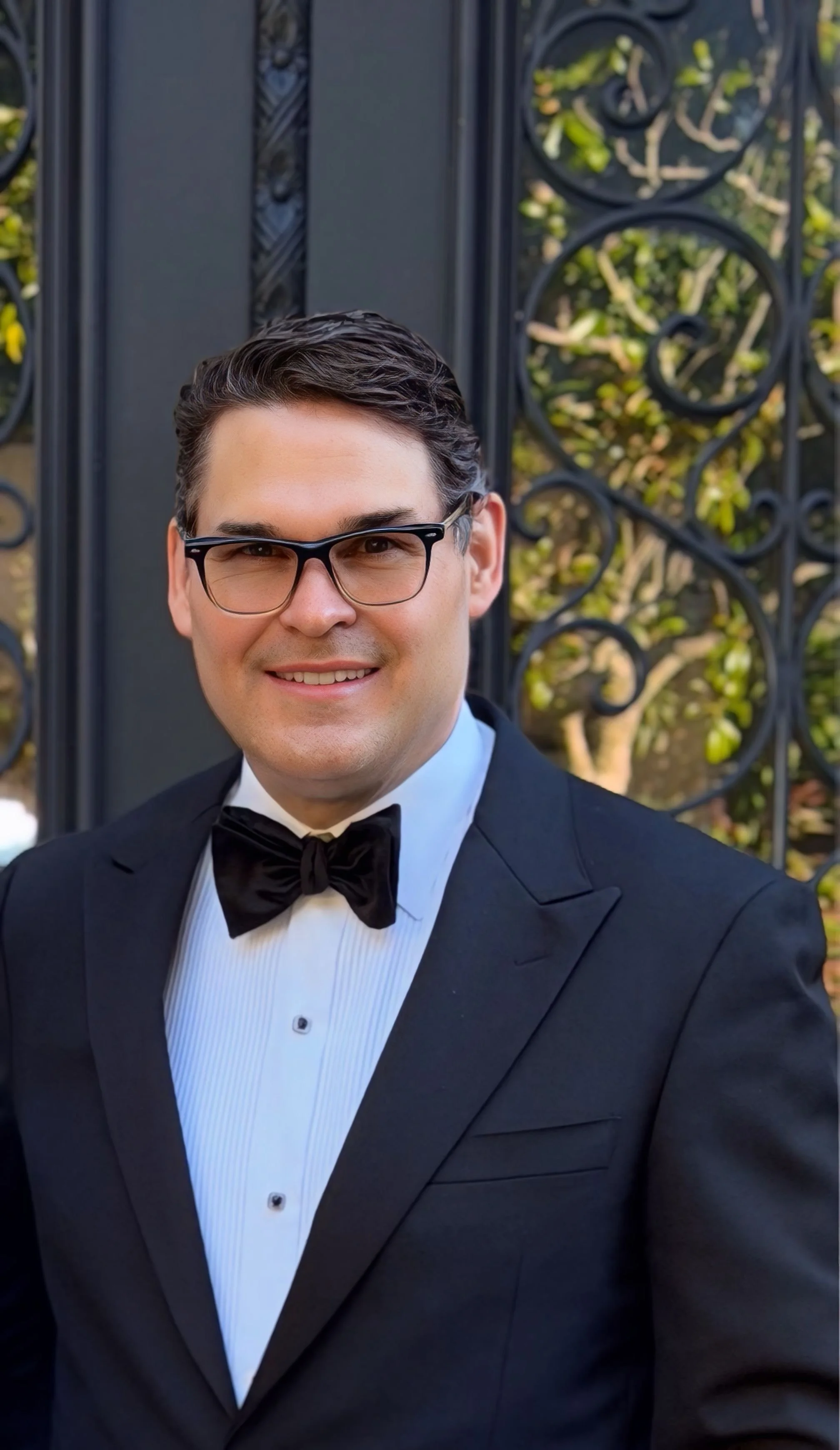 A man dressed in a tuxedo with a bowtie, wearing glasses, smiling outdoors in front of a decorative black wrought iron fence with trees and foliage in the background.