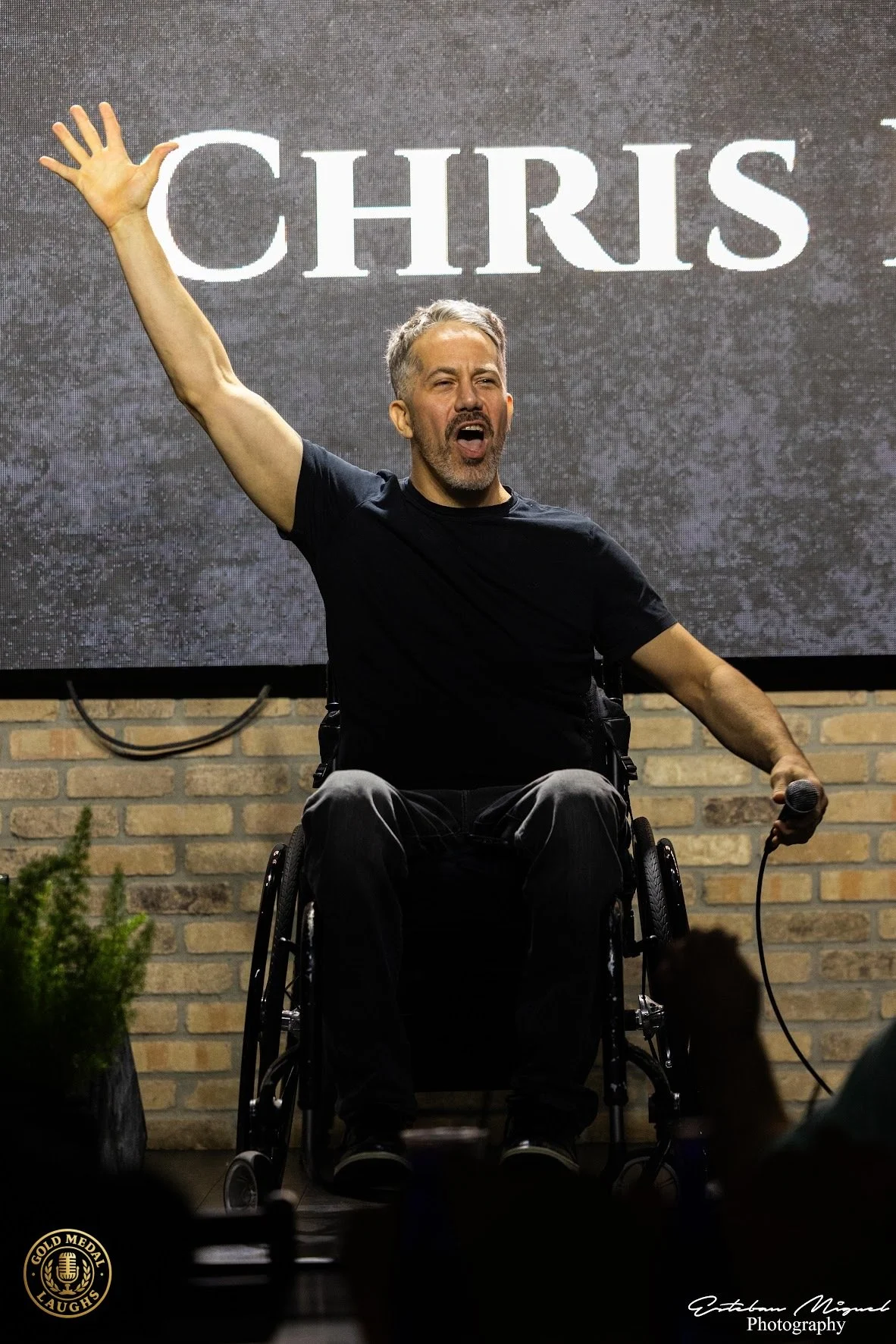 A man in a wheelchair holding a microphone in his right hand, raising his left arm, and gesturing with an open hand while speaking on stage. Behind him, a large screen displays the name 'CHRIS.' The background features a brick wall. The man appears t