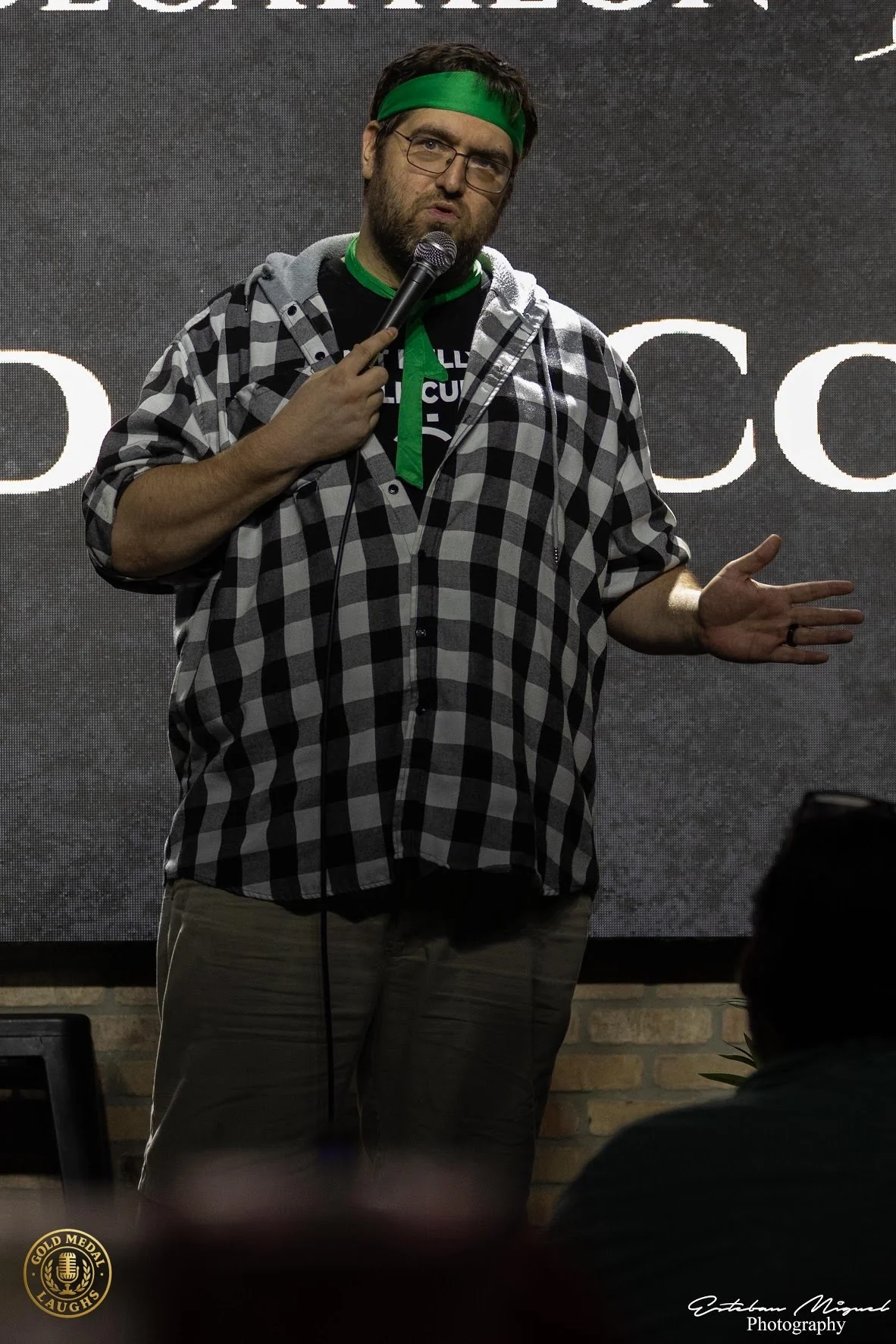 A man with a beard and glasses, wearing a green headband and a black T-shirt with white text, stands in front of a dark background with large white letters. He is holding a microphone in his right hand, wearing a grey and black checkered shirt, and m
