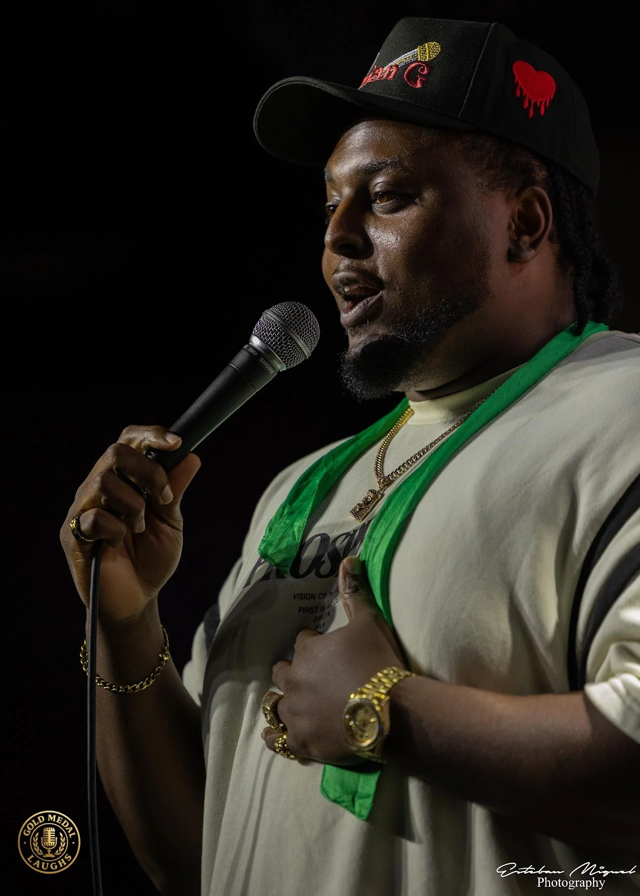 A man speaking into a microphone, wearing a black cap with red and yellow embroidered patches, a white shirt with green trim, and gold jewelry, against a dark background.