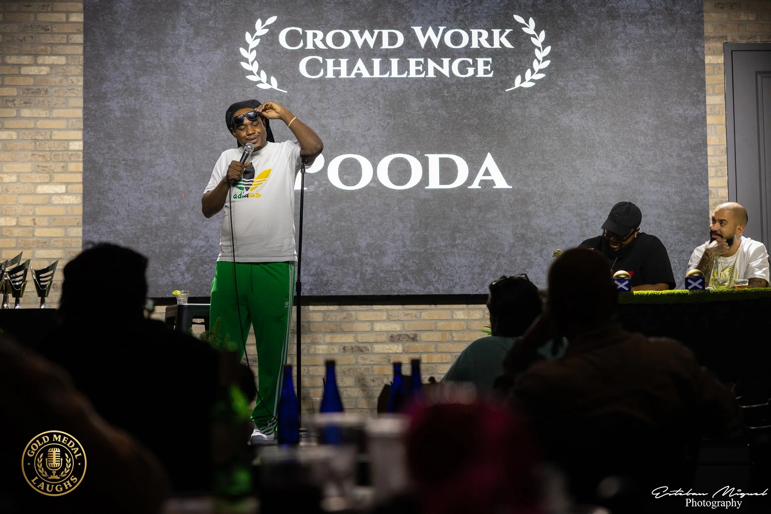 A man wearing sunglasses, a black head wrap, a white T-shirt, and green pants holds a microphone on stage during the Crowd Work Challenge event. Two men are seated at a table to his right, with one laughing and the other looking on. The background fe