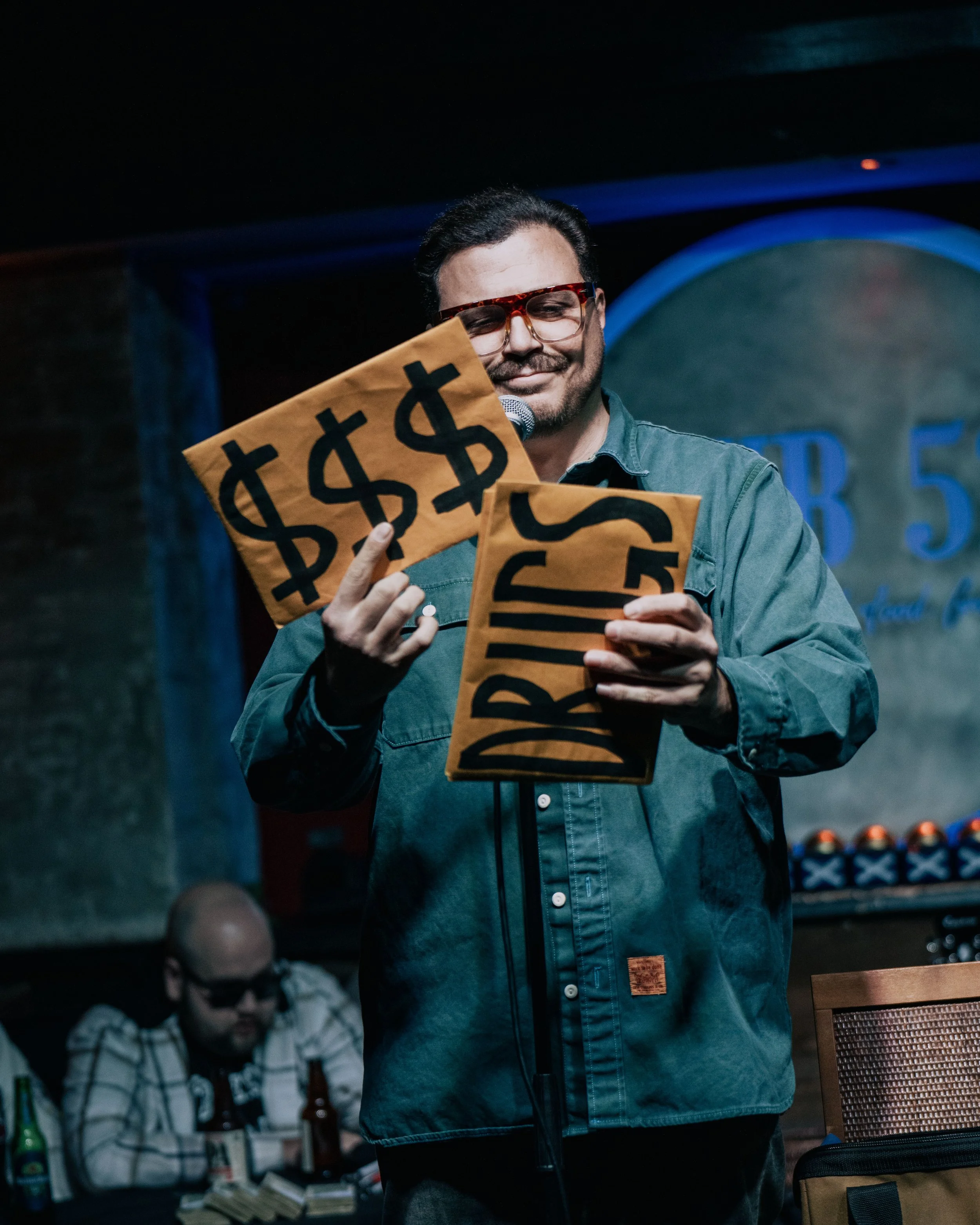 A man wearing sunglasses and a denim jacket is standing and smiling while holding some signs with dollar signs on them in a dimly lit room. Other people and objects are visible in the background.