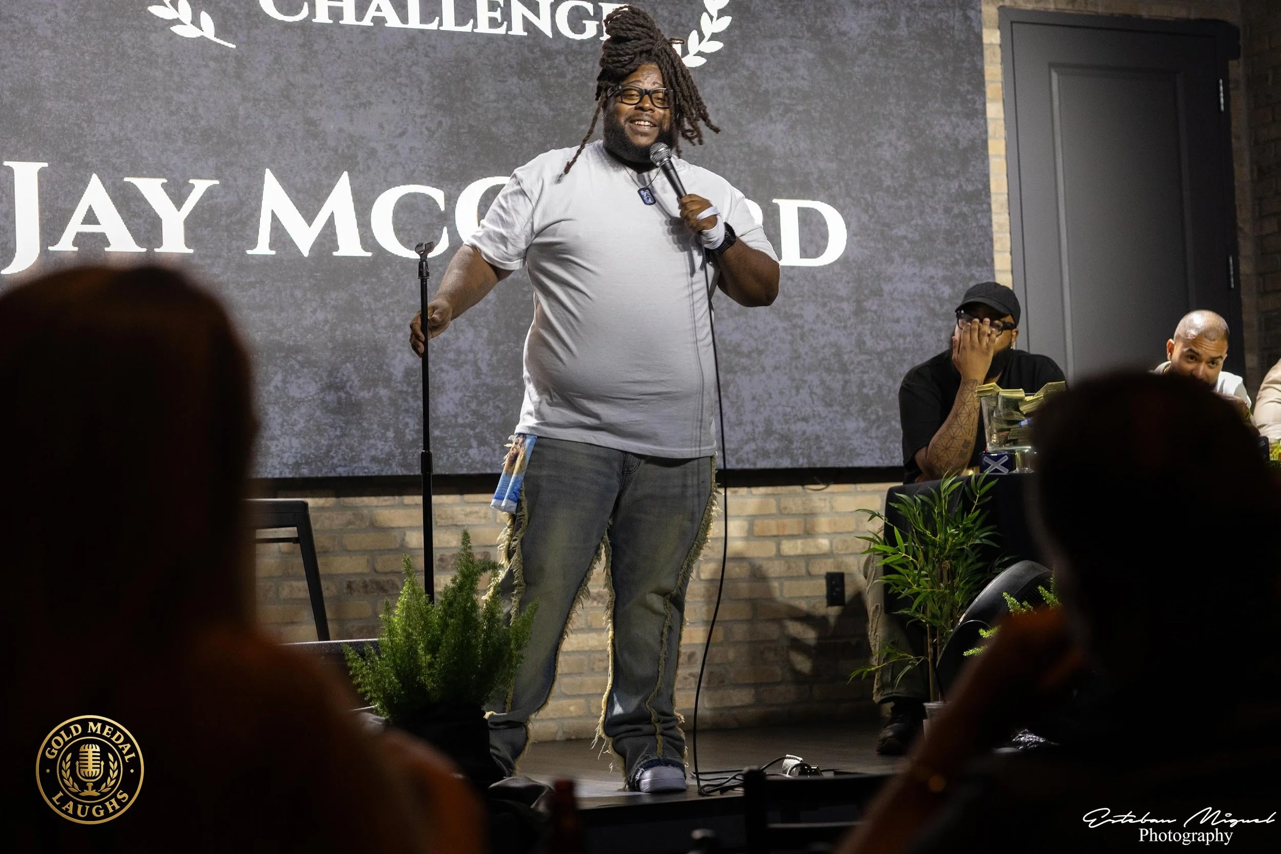 A man with dreadlocks, glasses, and a beard is speaking into a microphone on stage at a comedy show or event. He is wearing a light gray T-shirt, jeans with patches, and has a watch on his left wrist. Behind him is a large screen with the words 'CHAL