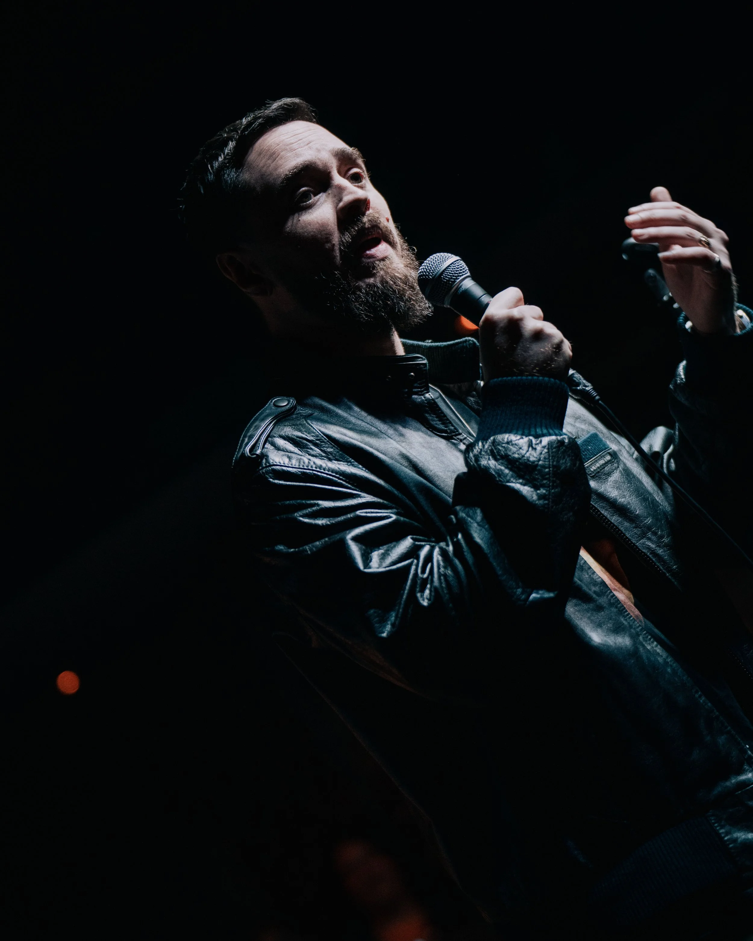 A bearded man is speaking into a microphone on a dark stage, wearing a leather jacket.