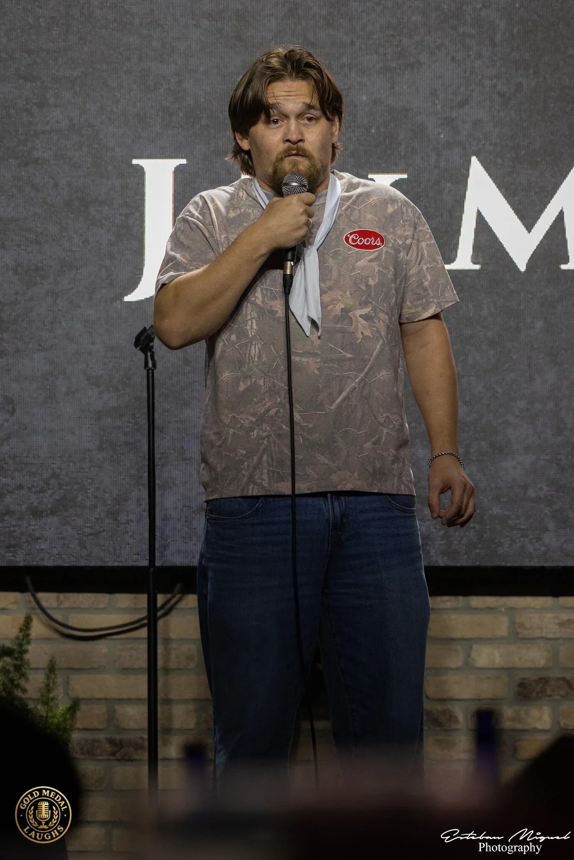 A man with medium-length brown hair, beard, wearing a camouflage shirt with a Coors patch, holds a microphone and stands in front of a screen with partially visible text, possibly at a comedy or entertainment event.