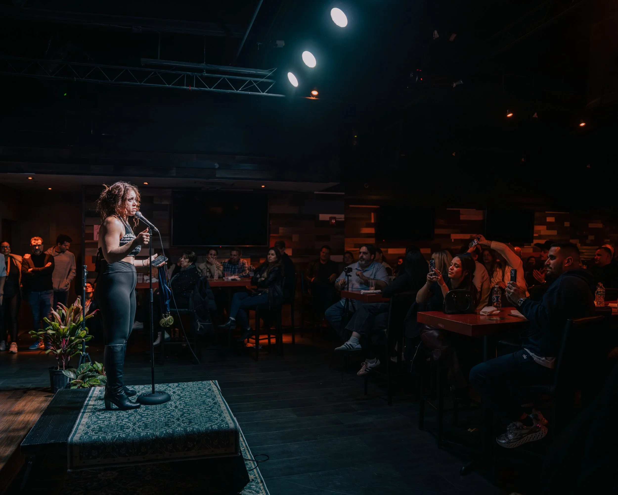 A woman with curly hair stands on a small stage with a microphone, speaking or performing in front of an audience in a dimly lit room with a dark wooden interior and colorful lighting. The audience, composed of multiple people, is seated at tables wa