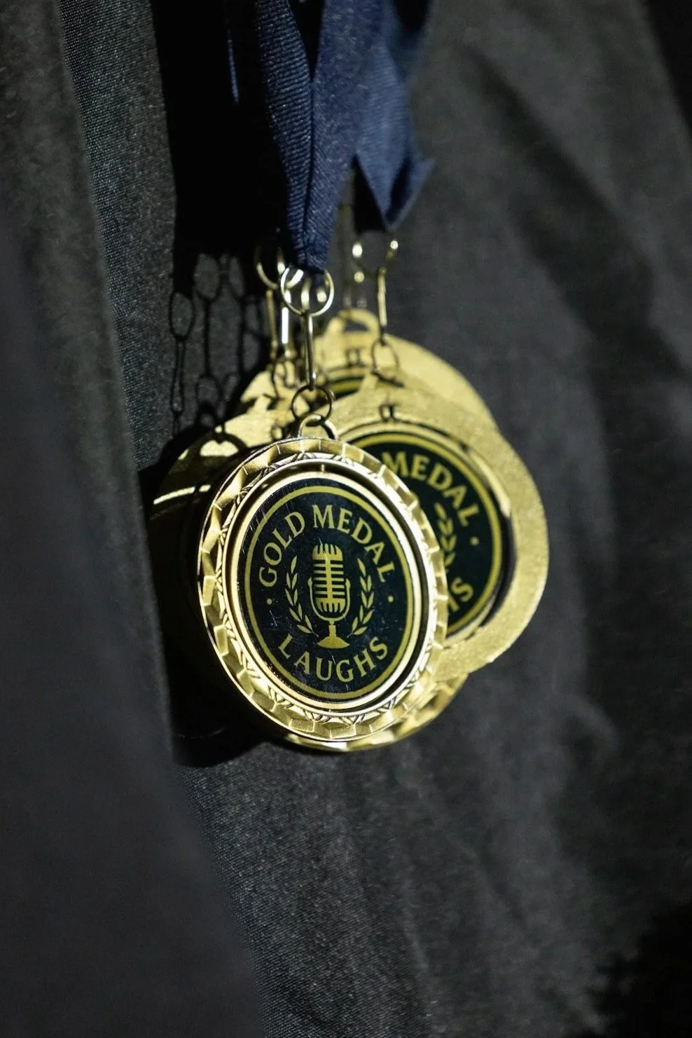 Close-up of gold medals hanging from a dark fabric, each with a circular design featuring a microphone and the text 'Gold Medal Laughs'.