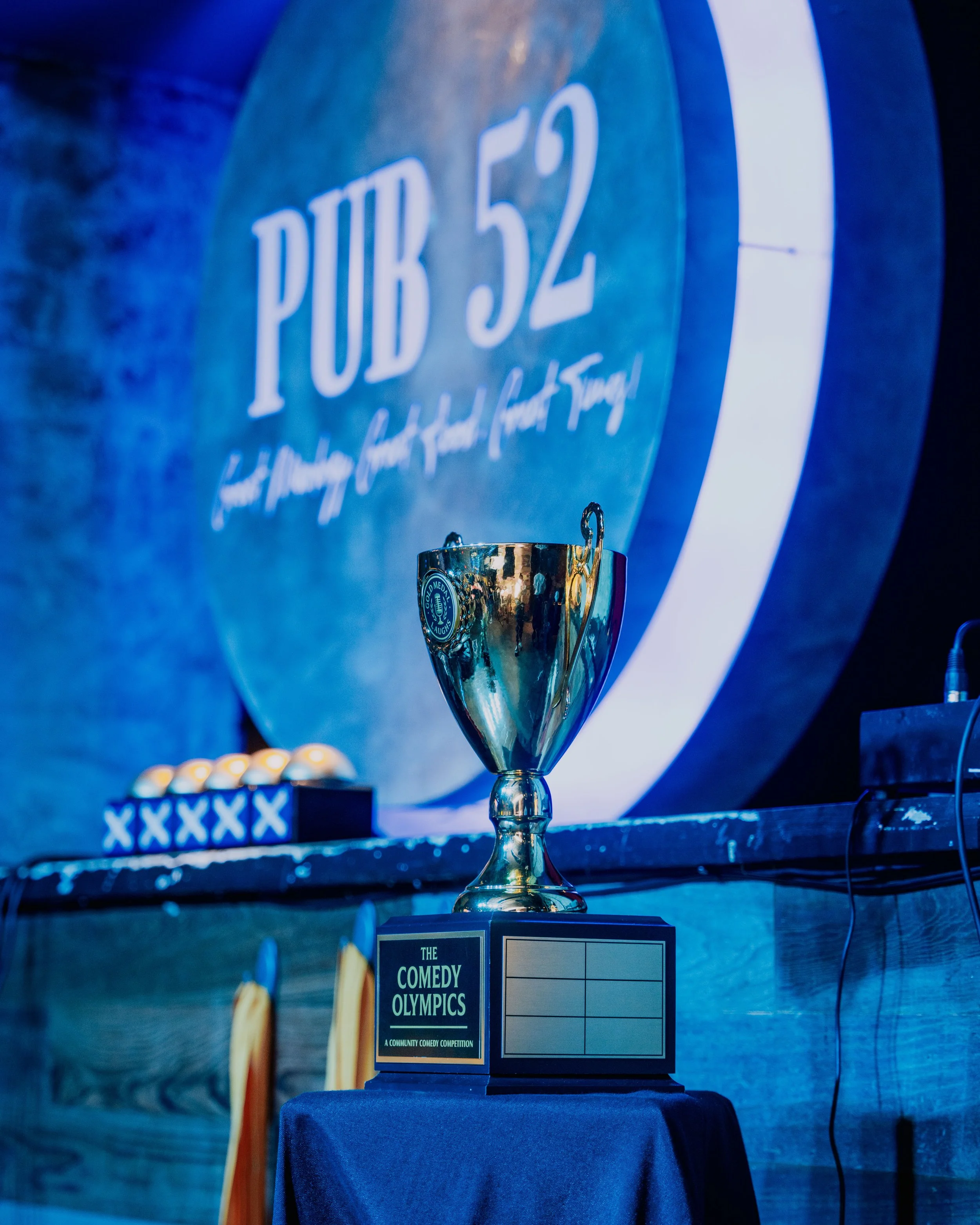 A trophy on a table with a sign that reads "The Comedy Olympics: A Community Comedy Competition" in front of a blue background featuring a large sign that says "PUD 52".