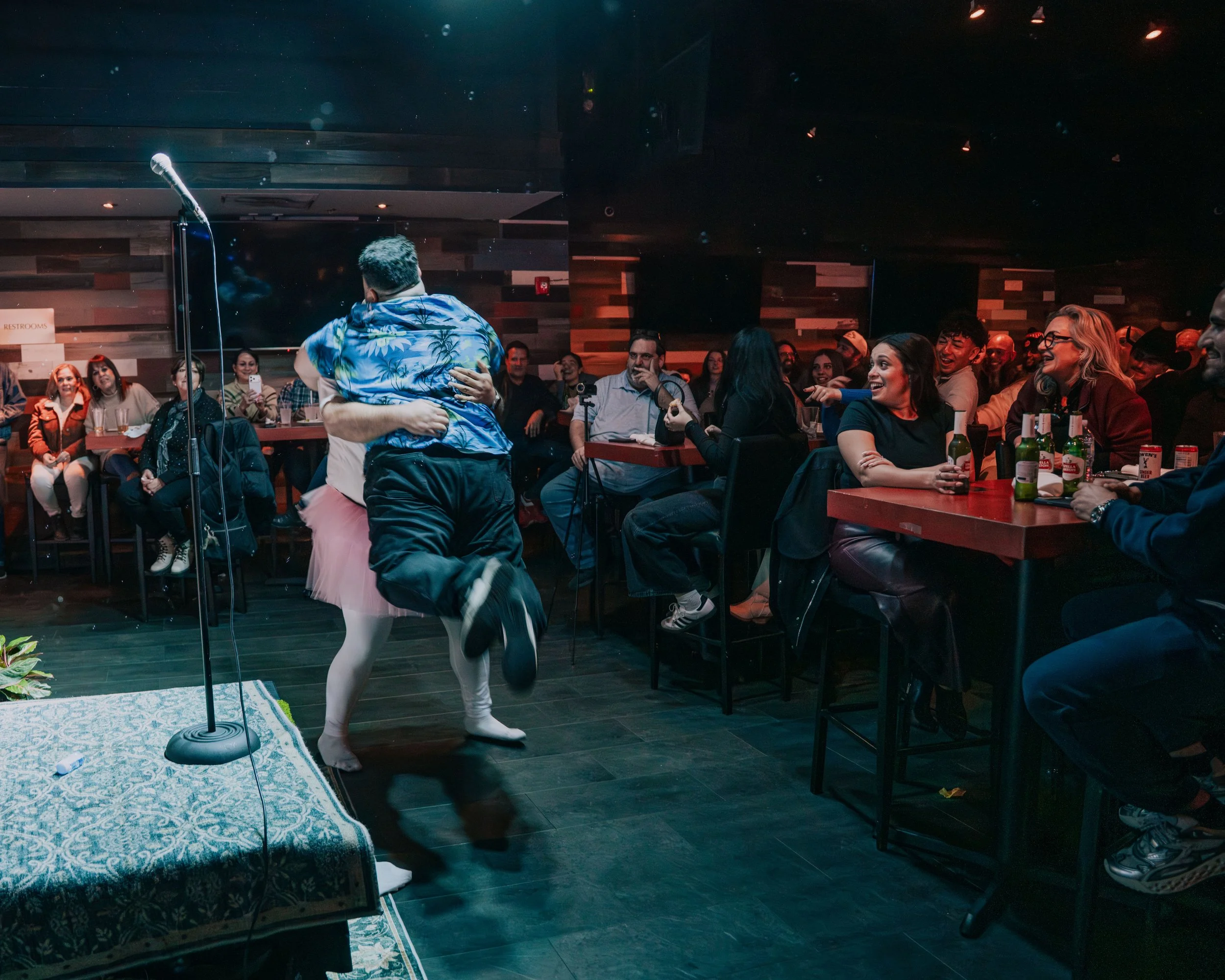 A man and woman embracing on stage at a comedy show, with an audience laughing and clapping in a dimly lit venue.