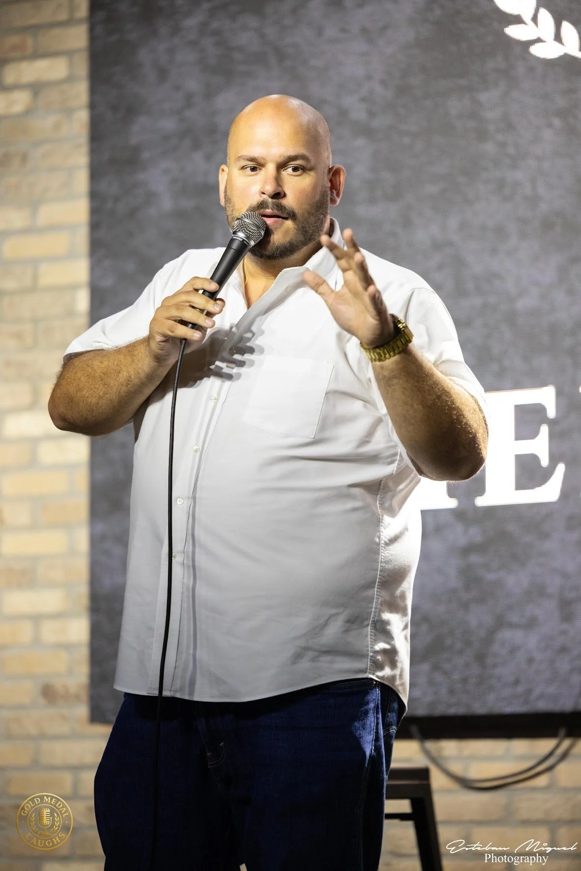 A man speaking into a microphone on stage, wearing a white shirt and dark pants, with a brick wall and a partial sign in the background.