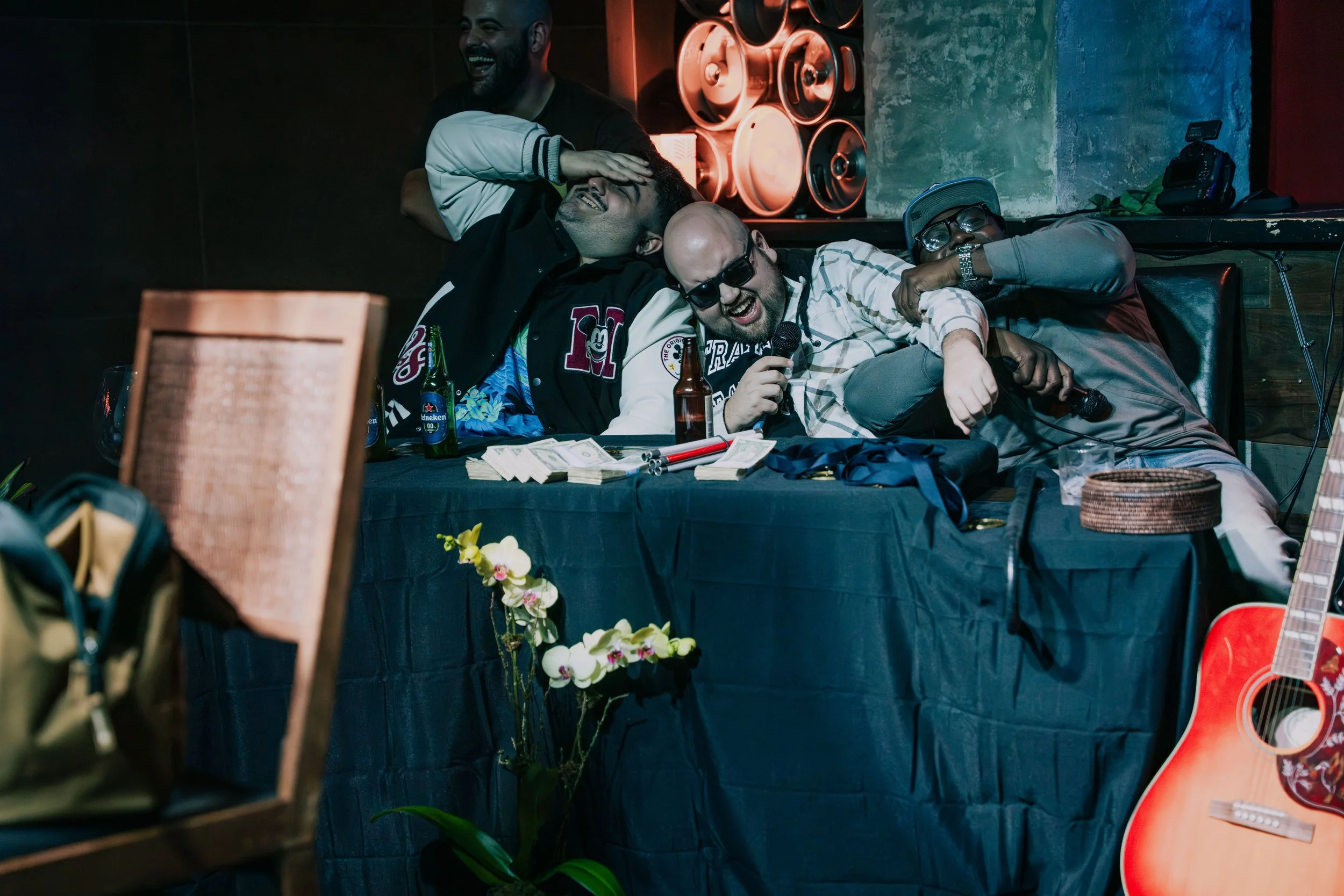 Group of four friends at a bar, laughing and enjoying each other's company. One person is holding a microphone, with cash on the table, beer bottles, and a guitar in the foreground.