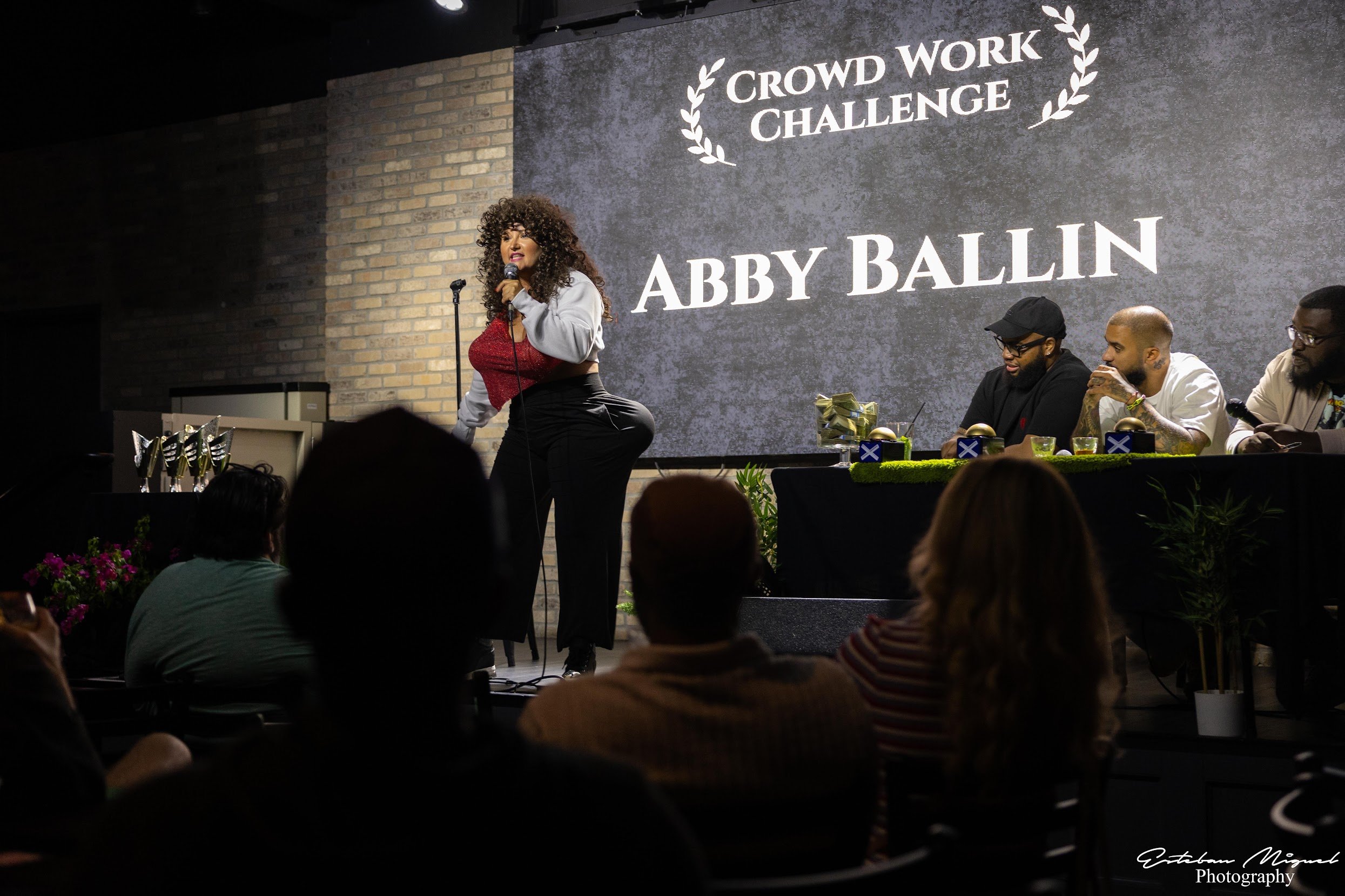 A woman with curly hair wearing a red top and gray jacket speaking into a microphone on stage during a crowd work challenge event. Behind her is a large screen displaying the event name 'Crowd Work Challenge' and the name 'Abby Ballin'. Four men are 