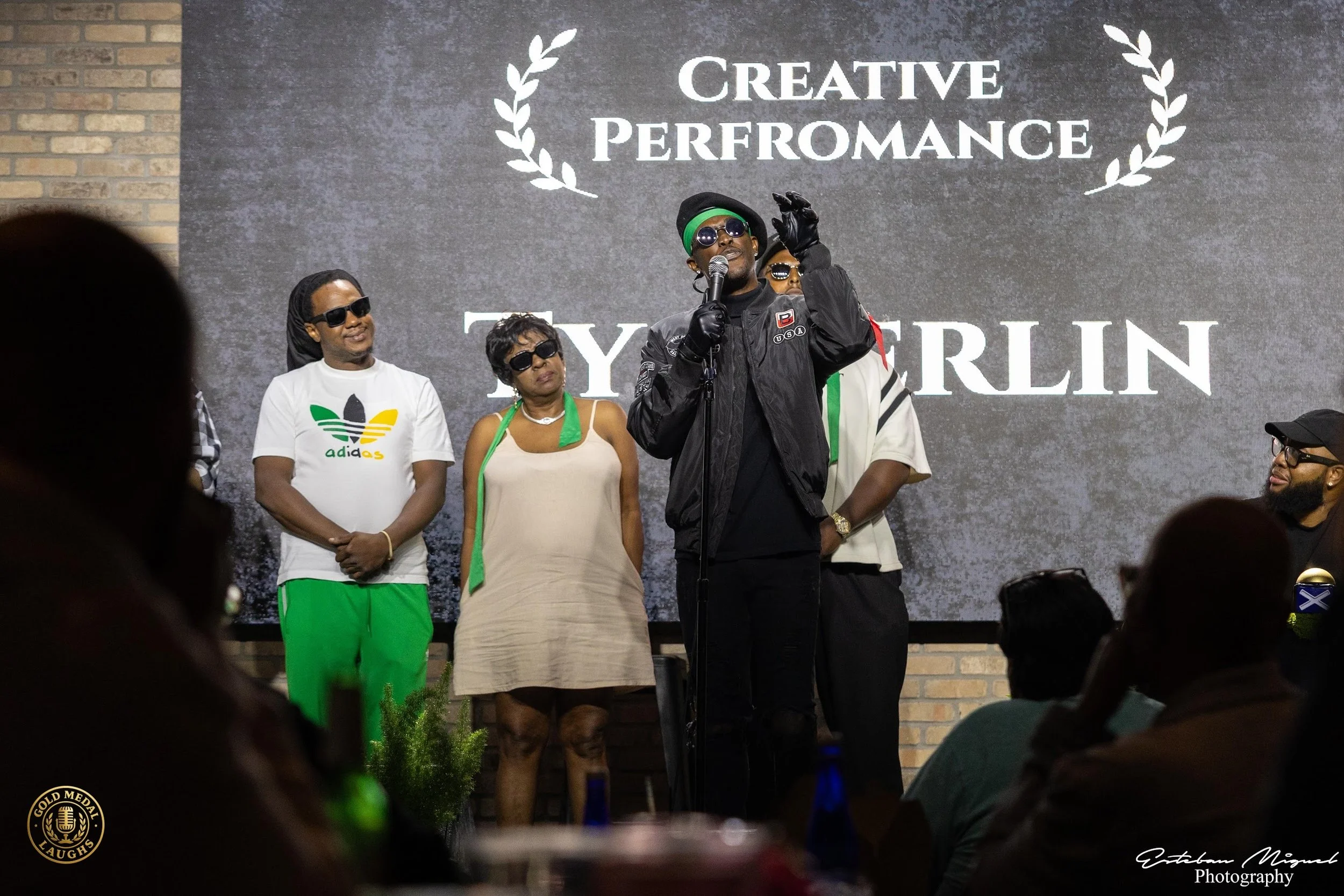 A group of people standing on stage under a sign that reads 'Creative Performance by Tyrellin'. The man at the center, wearing sunglasses, a black jacket, and gloves, is speaking into a microphone. Women and men surround him, some wearing sunglasses,