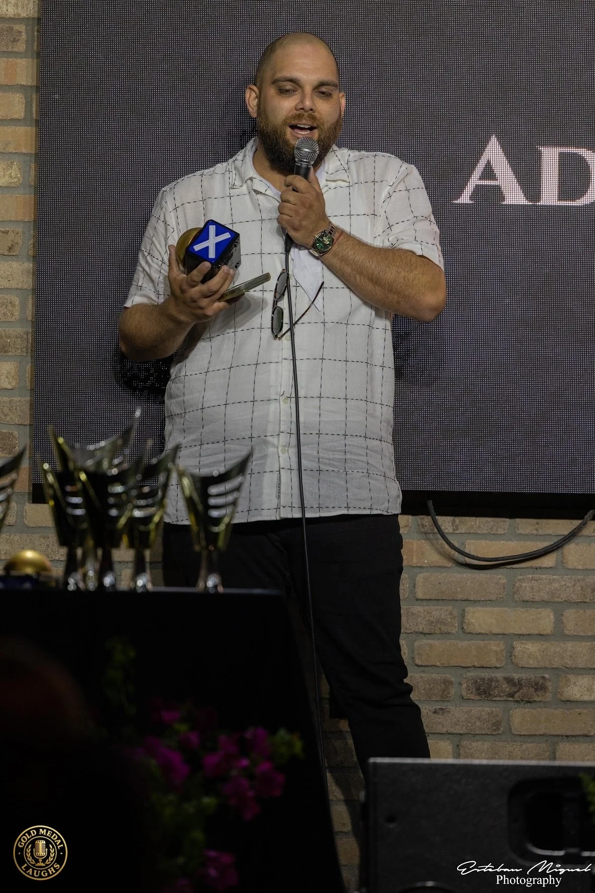 A man holding a microphone and a plaque with a blue and white star symbol on it, standing in front of a black digital screen displaying the text 'AD'. He is wearing a white shirt with black checkered lines, a wristwatch, and glasses hanging from his 