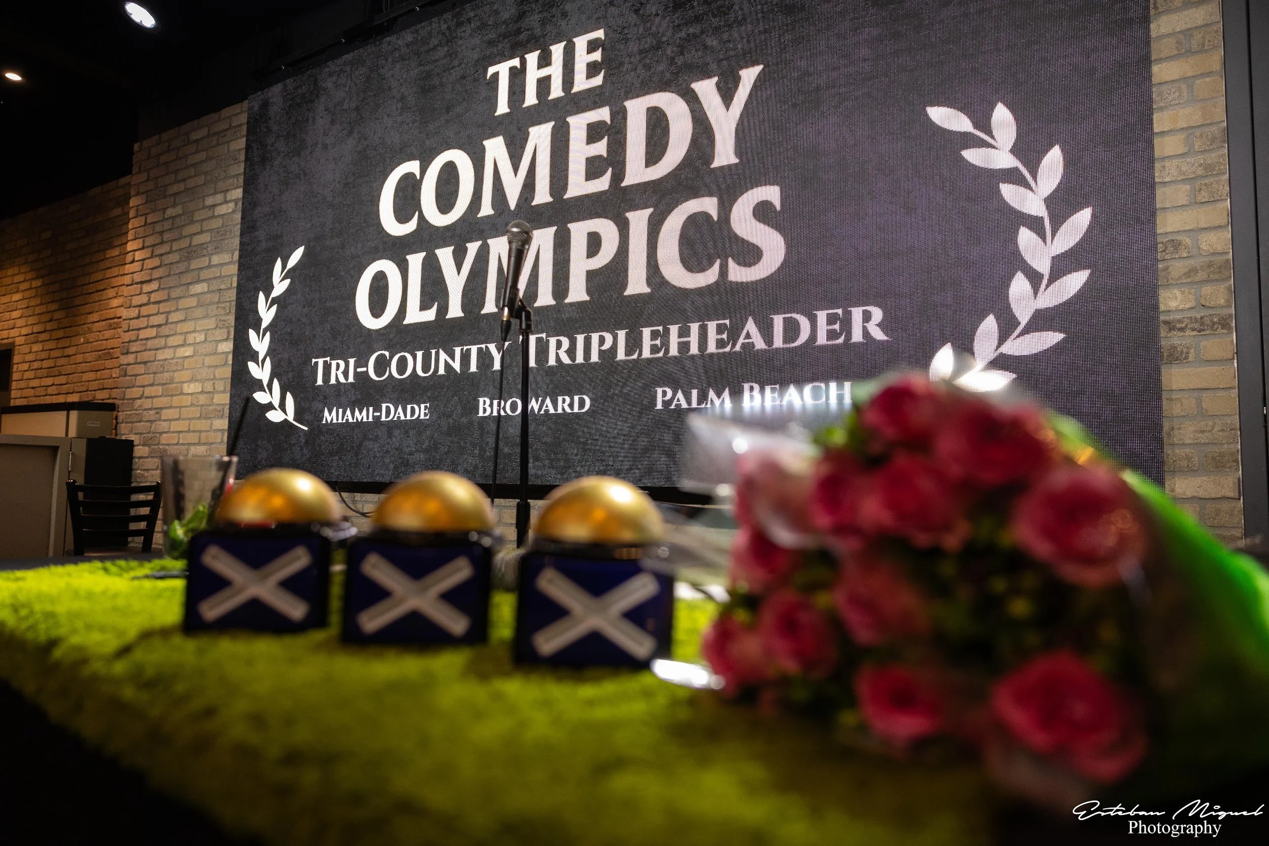 Stage setup for The Comedy Olympics event, featuring a large digital screen with event details, a microphone on stand, and decorations with flowers and medals in the foreground.