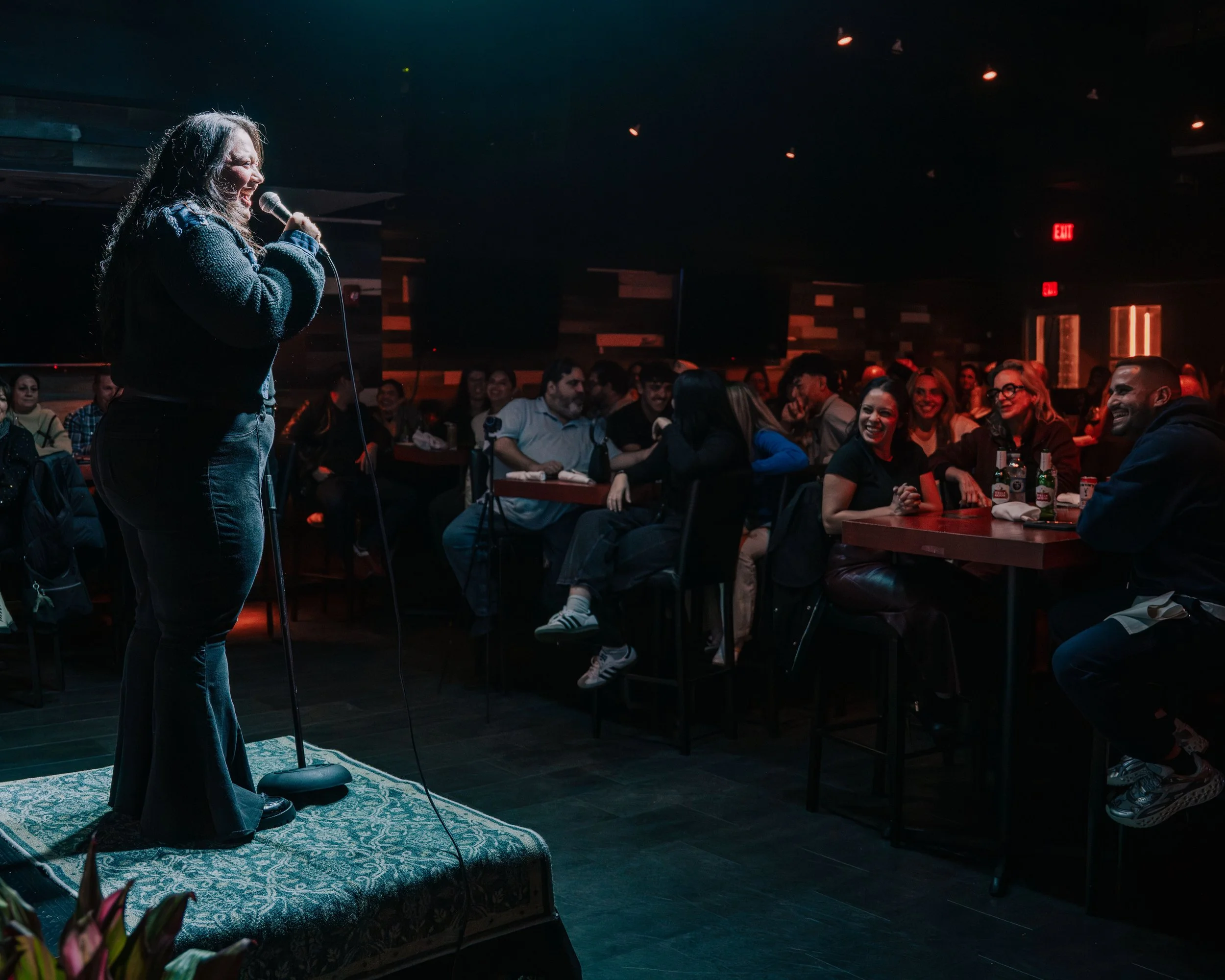 A woman performing stand-up comedy on stage in front of an audience at a comedy club. The audience is laughing and attentively watching her.