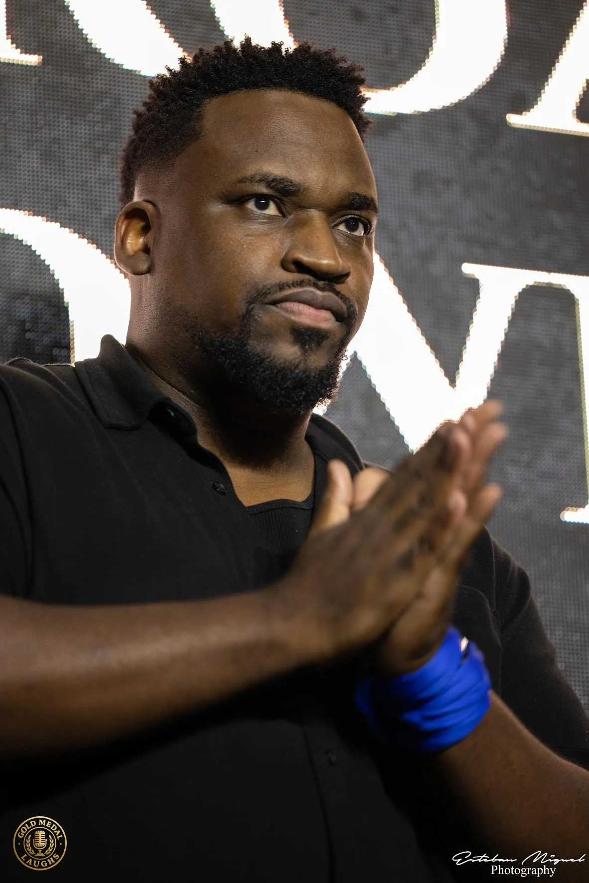 A man with short curly hair and a beard poses with his hands pressed together in front of a lit sign background. He is wearing a black shirt and a blue wristband, and appears to be at a promotional or entertainment event.