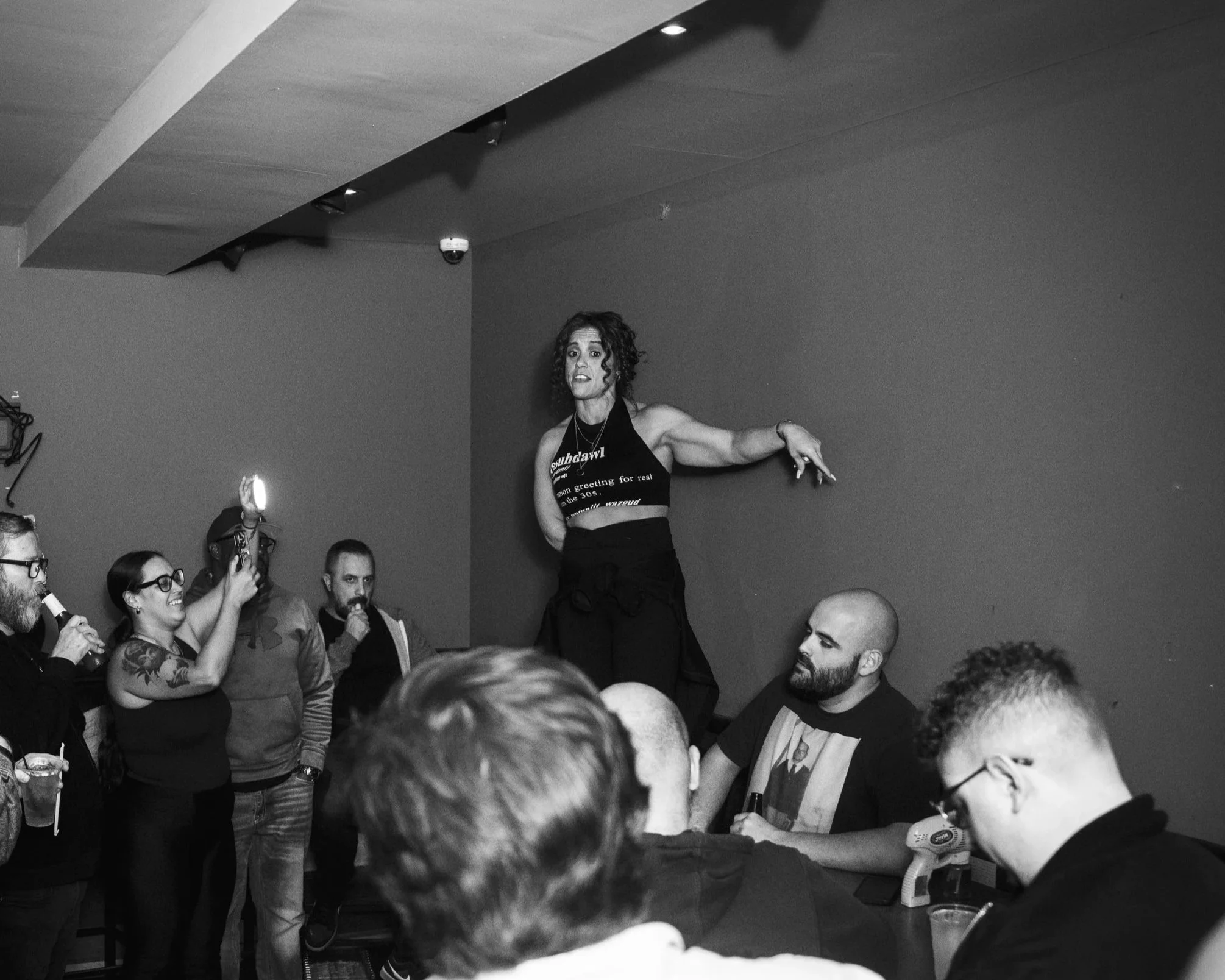 A woman with dark, curly hair stands on a platform in front of a crowd, gesturing with her right arm extended and her left arm bent. The audience, mostly adults, are watching and some are capturing the moment with their phones. The setting appears to be indoors, possibly at an event or presentation.