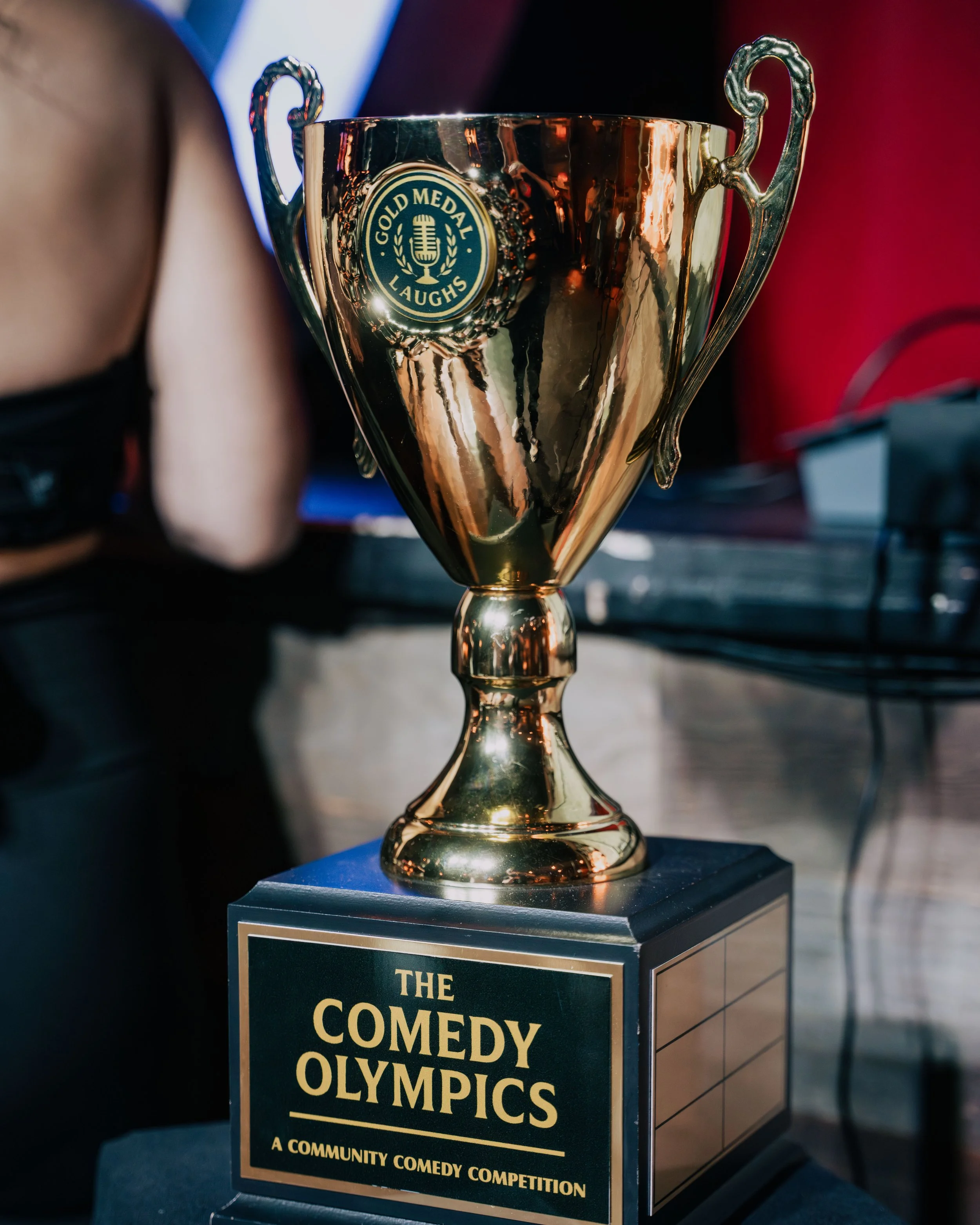 Golden trophy with handles, inscribed with 'Gold Medal Laughs' and a microphone logo, placed on a podium at the Community Comedy Olympics event.
